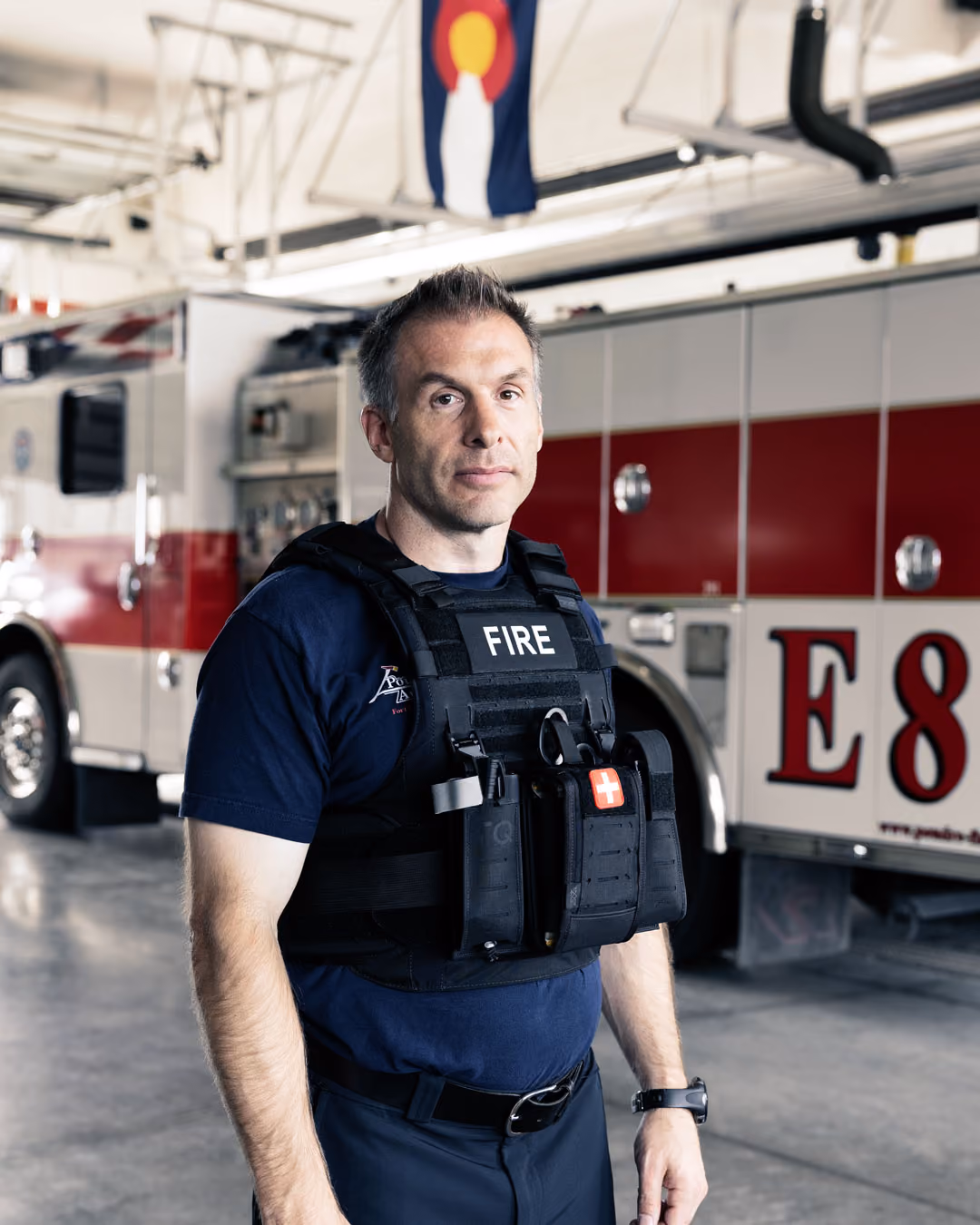 Firefighter wearing tactical vest standing in front of a fire engine inside a fire station.