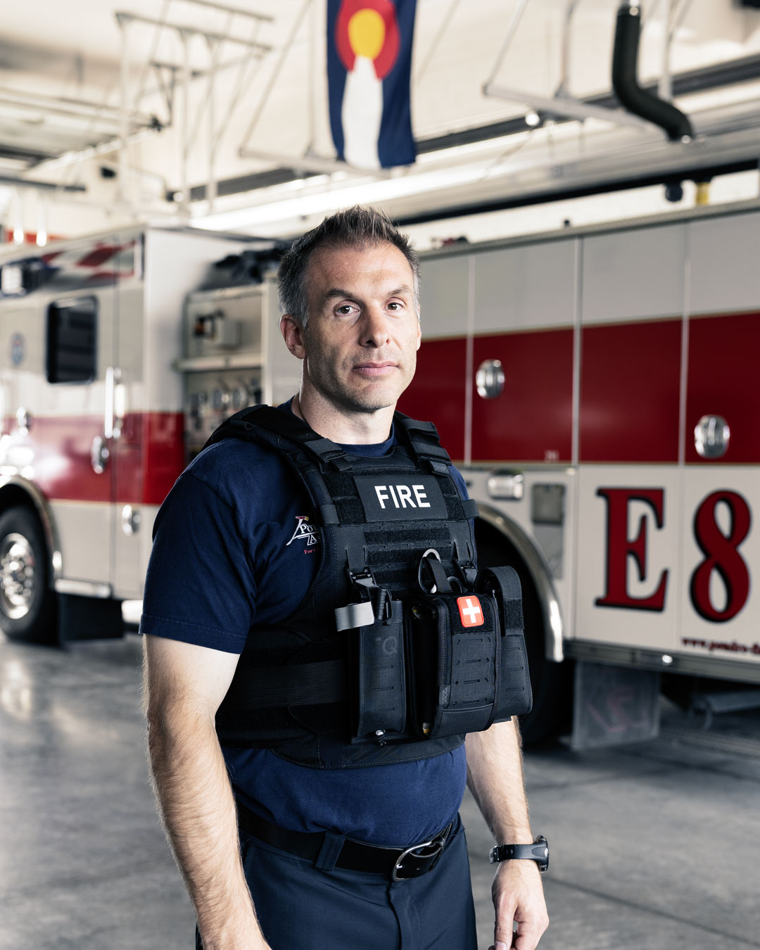 Firefighter wearing tactical vest standing in front of a fire engine inside a fire station.