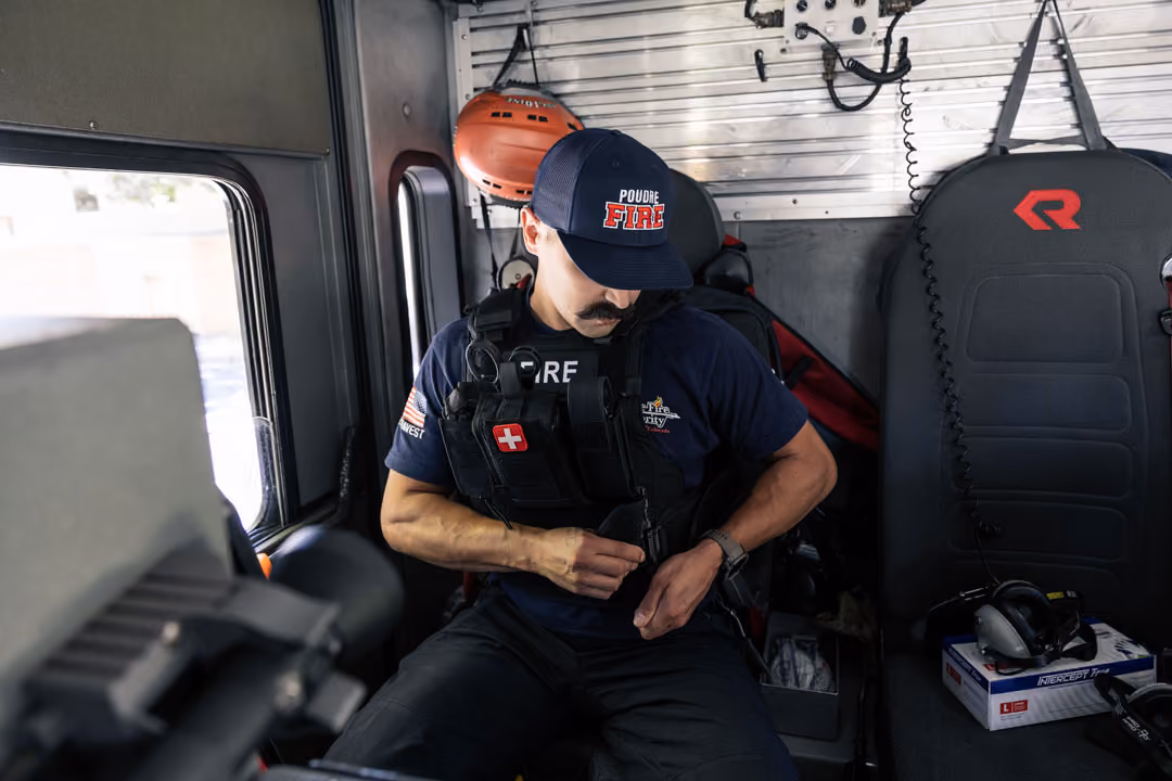 Firefighter inside vehicle adjusting gear vest, wearing navy shirt and Poudre Fire hat.