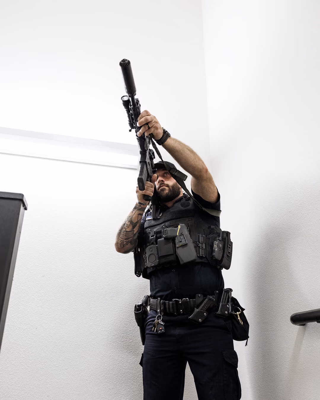 Man in tactical gear aiming a rifle upwards while standing in a white stairwell.