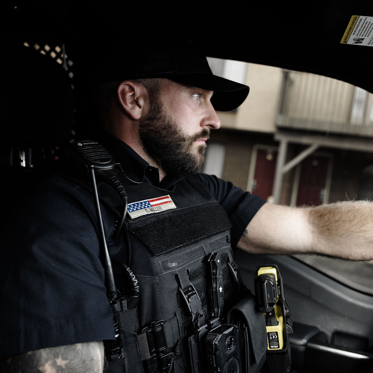 Bearded police officer in tactical vest driving a vehicle with focused expression.