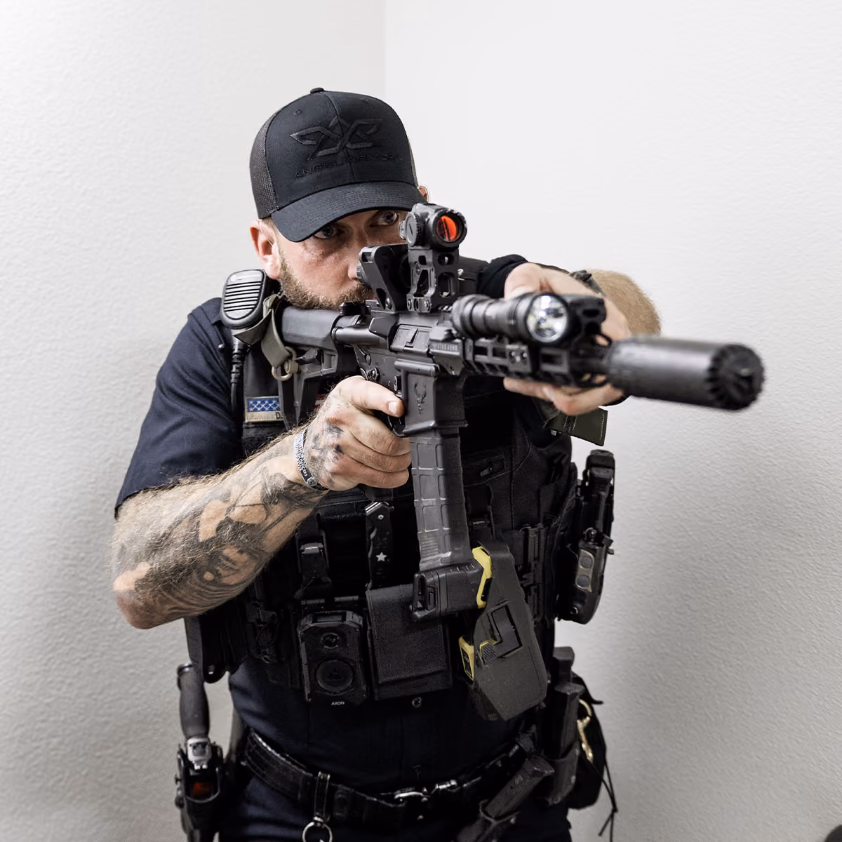 Tattooed law enforcement officer aiming a tactical rifle with attachments indoors.