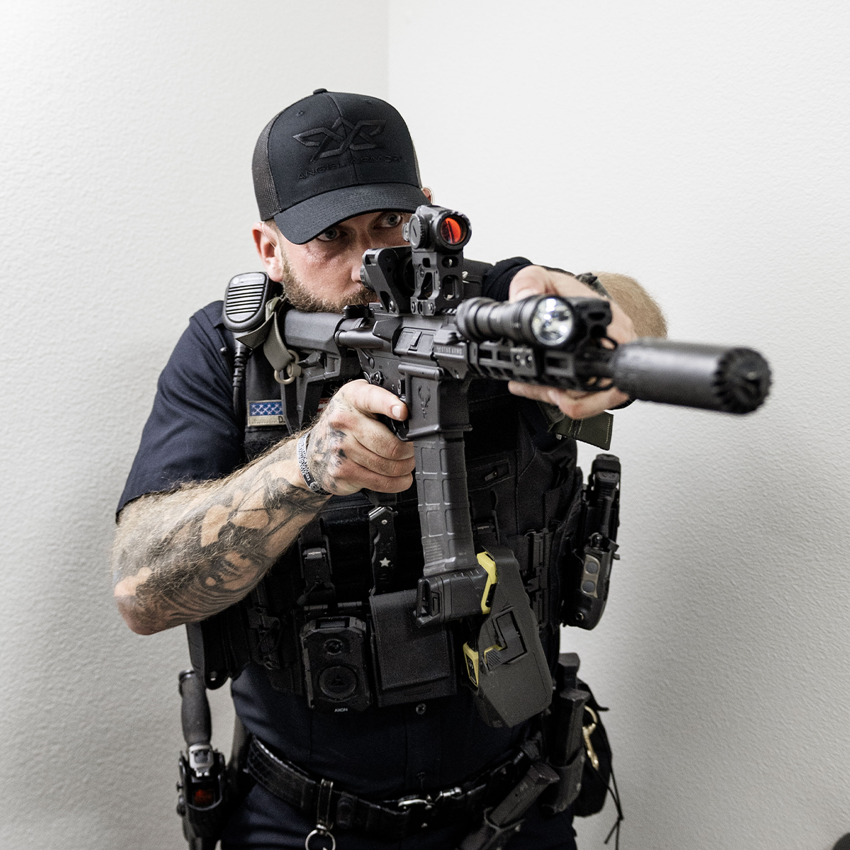 Tattooed law enforcement officer aiming a tactical rifle with attachments indoors.