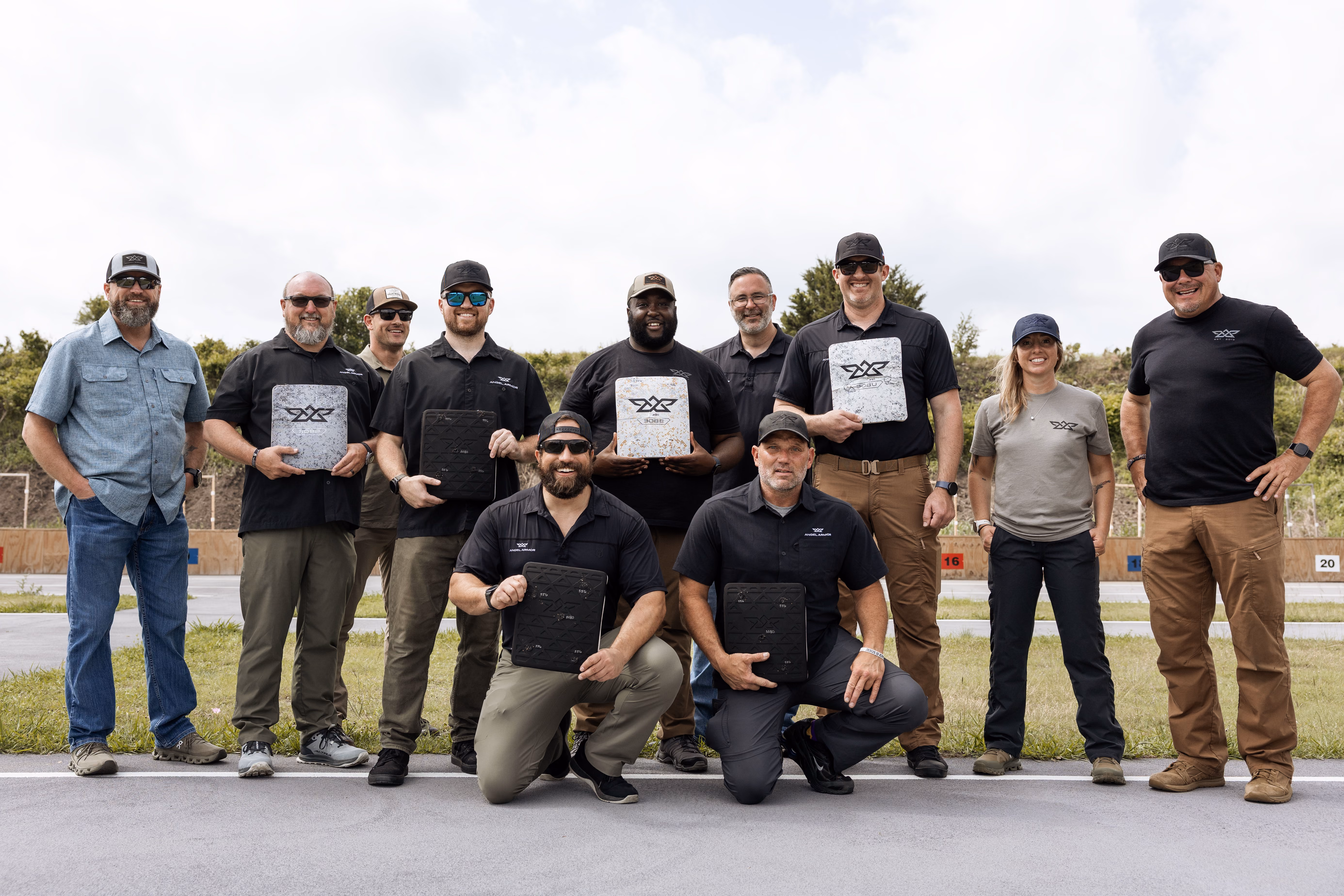 Group of eleven people posing outdoors, some holding protective shooting plates with logos.