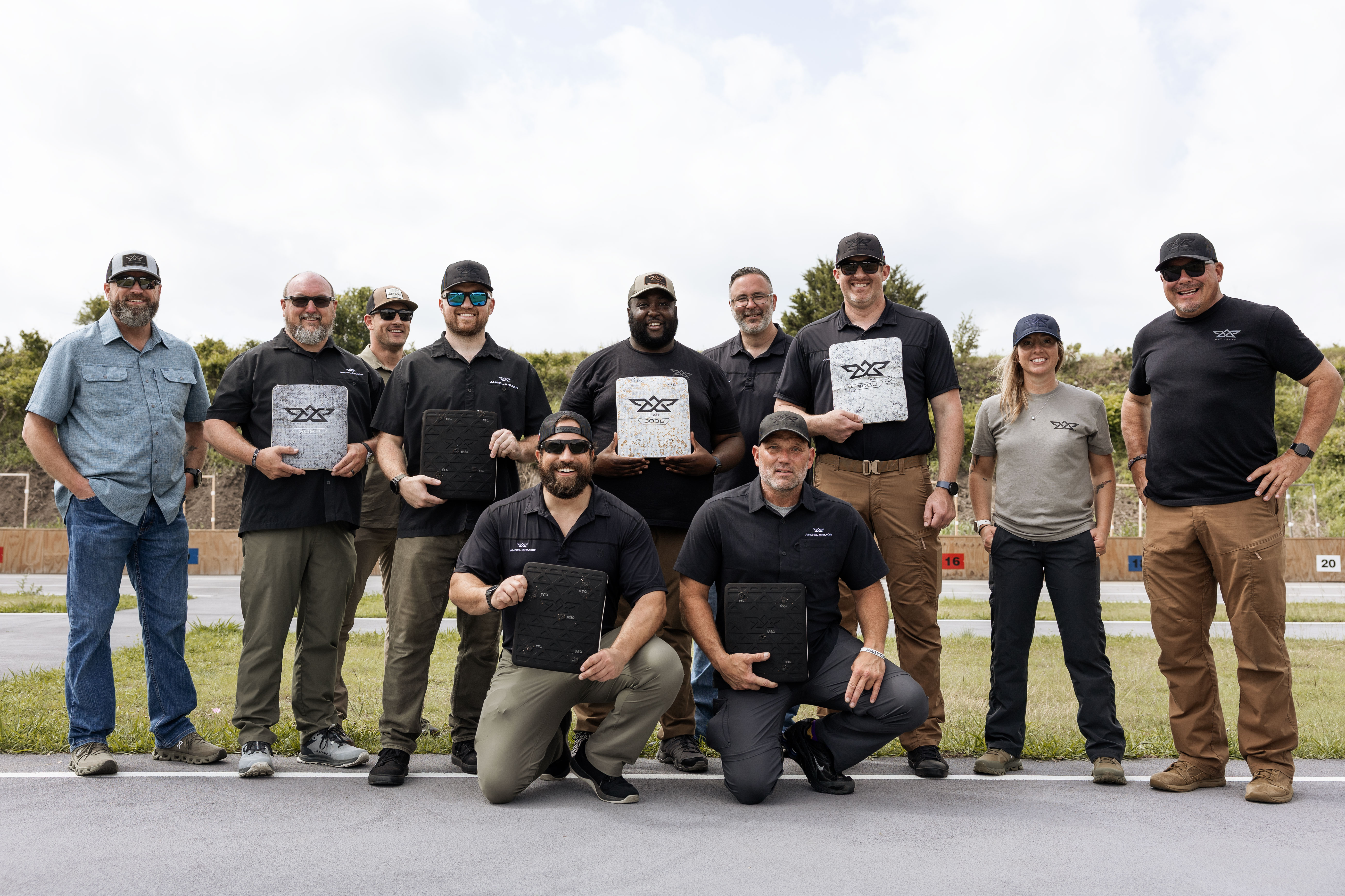 Group of eleven people posing outdoors, some holding protective shooting plates with logos.