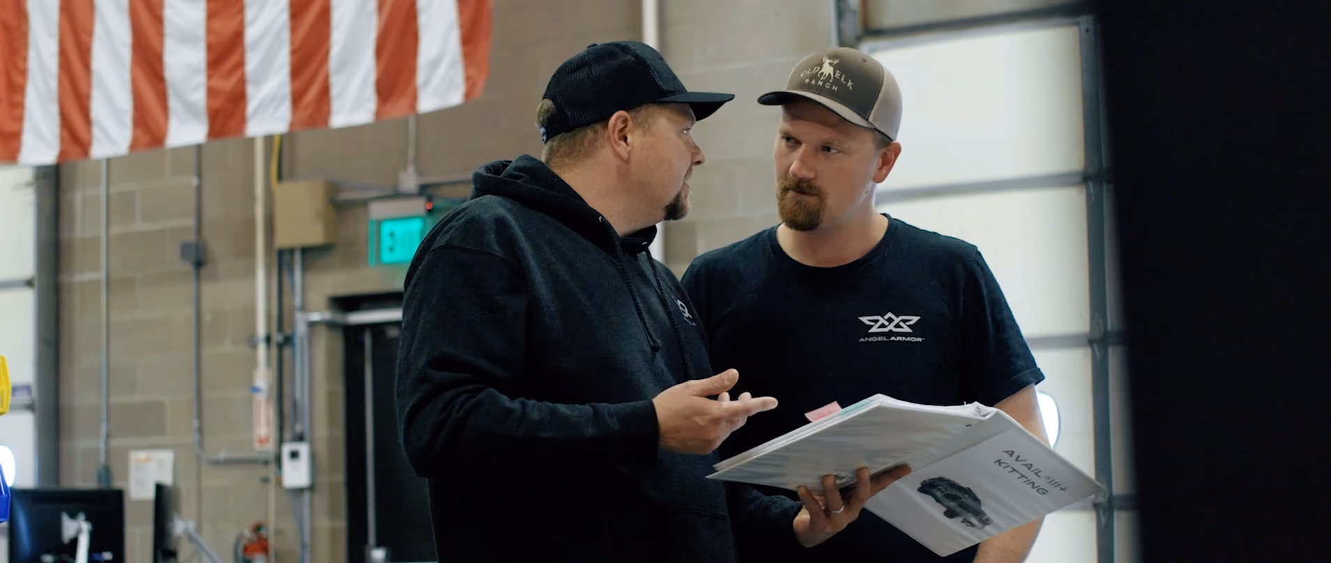 Two men in caps discuss while holding a binder inside a workshop with an American flag overhead.