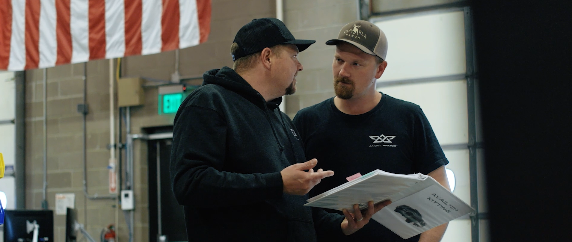 Two men in caps discuss while holding a binder inside a workshop with an American flag overhead.