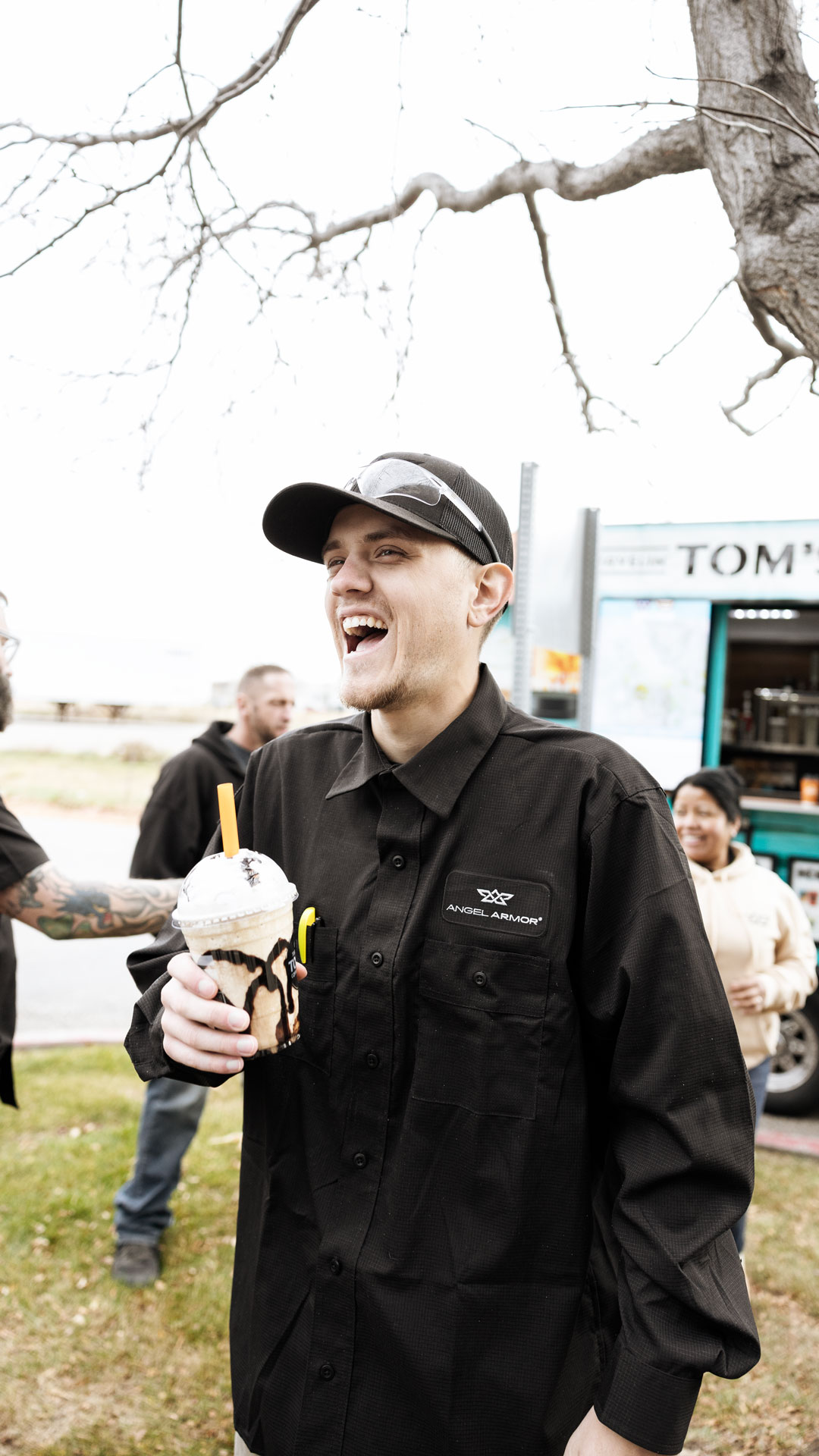 Man in black Angel Armor shirt smiling and holding a chocolate-drizzled iced coffee outdoors.
