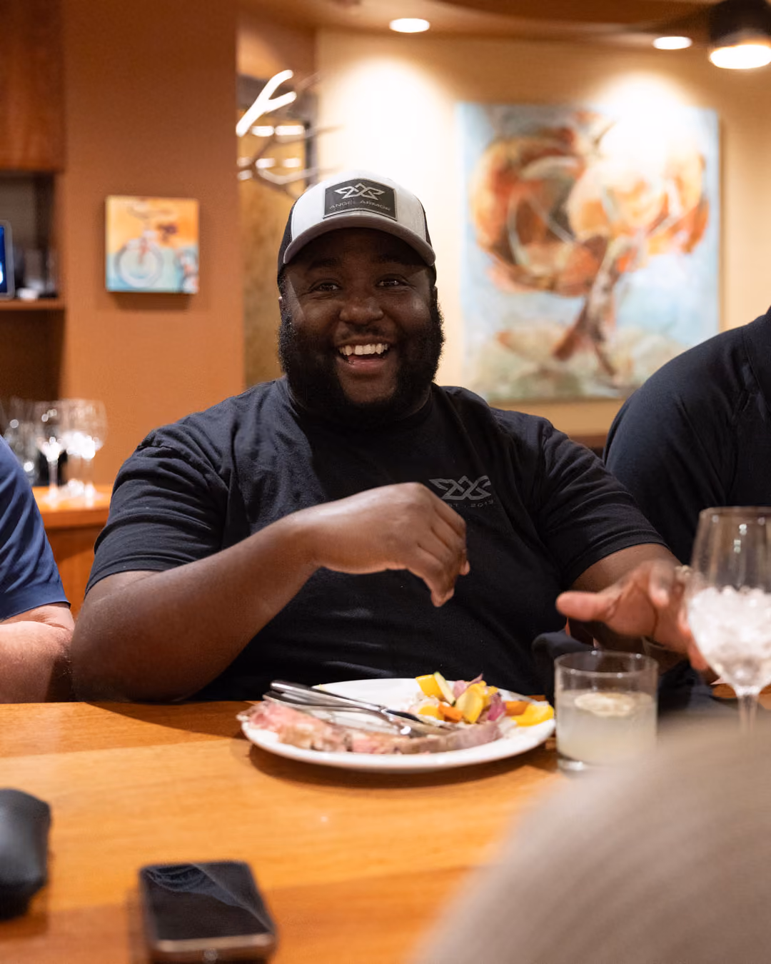 Smiling man in a cap sitting at a table with a plate of food and drinks in a warmly lit room.