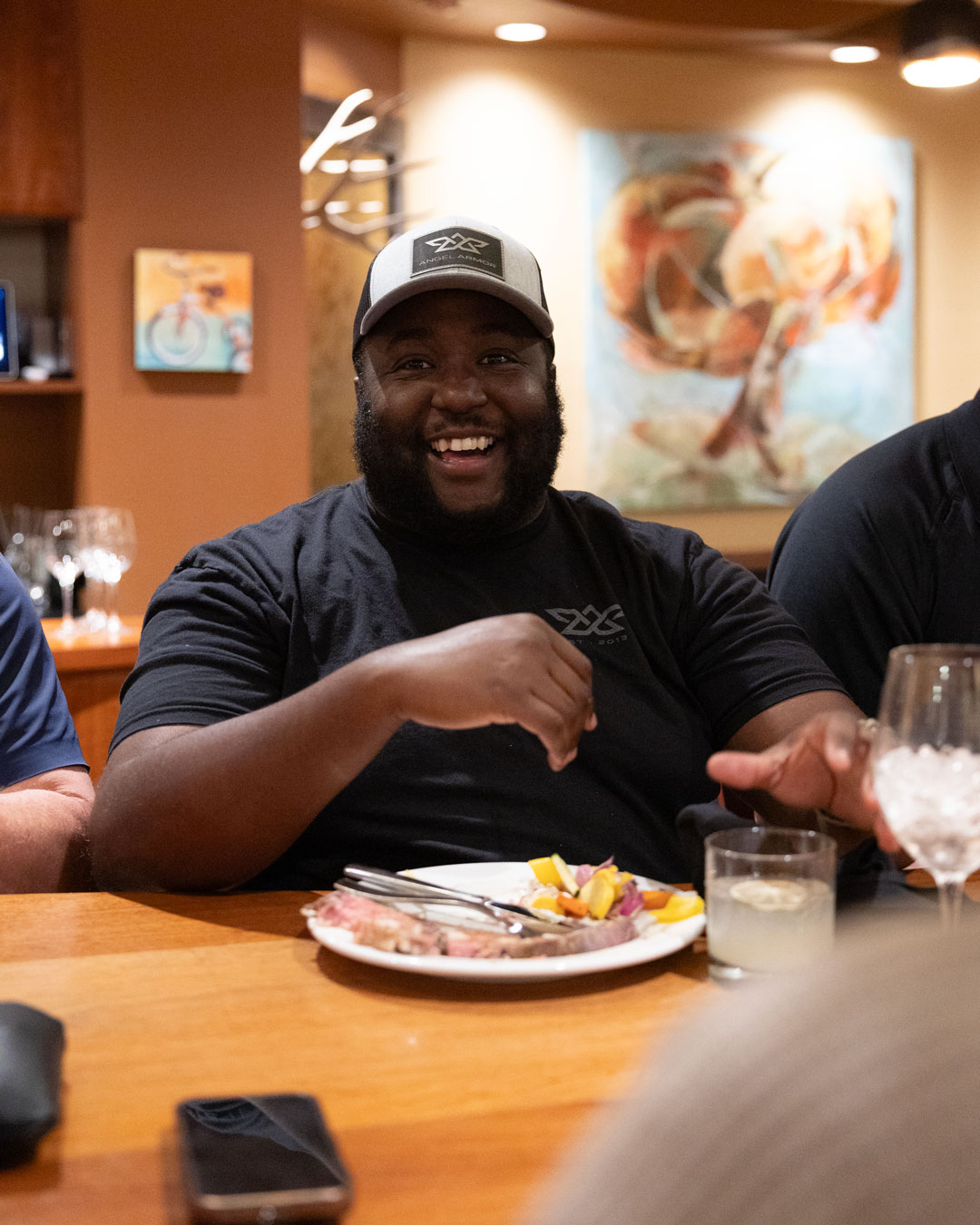 Smiling man in a cap sitting at a table with a plate of food and drinks in a warmly lit room.