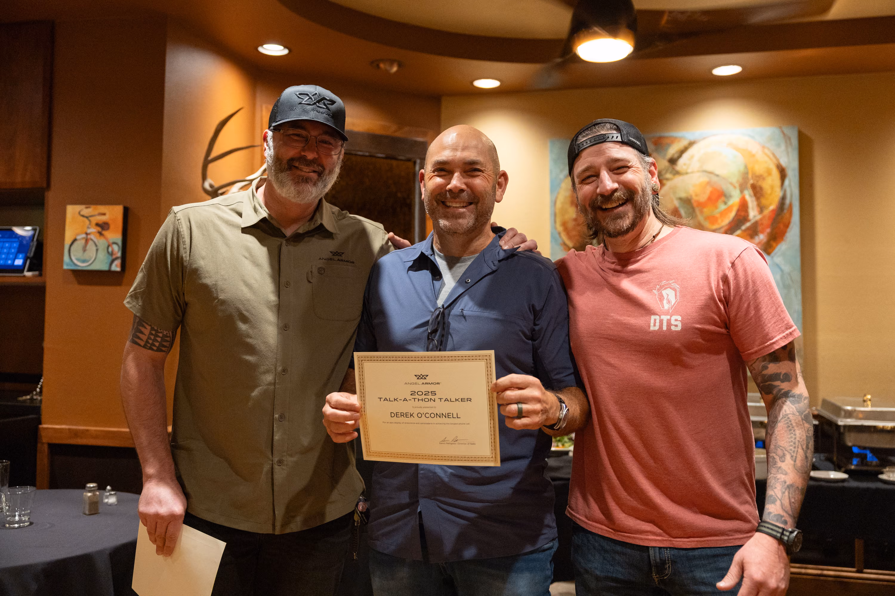 Three men standing indoors smiling, one holding a certificate recognizing Derek O'Connell for Talk-A-Thon Talker 2025.