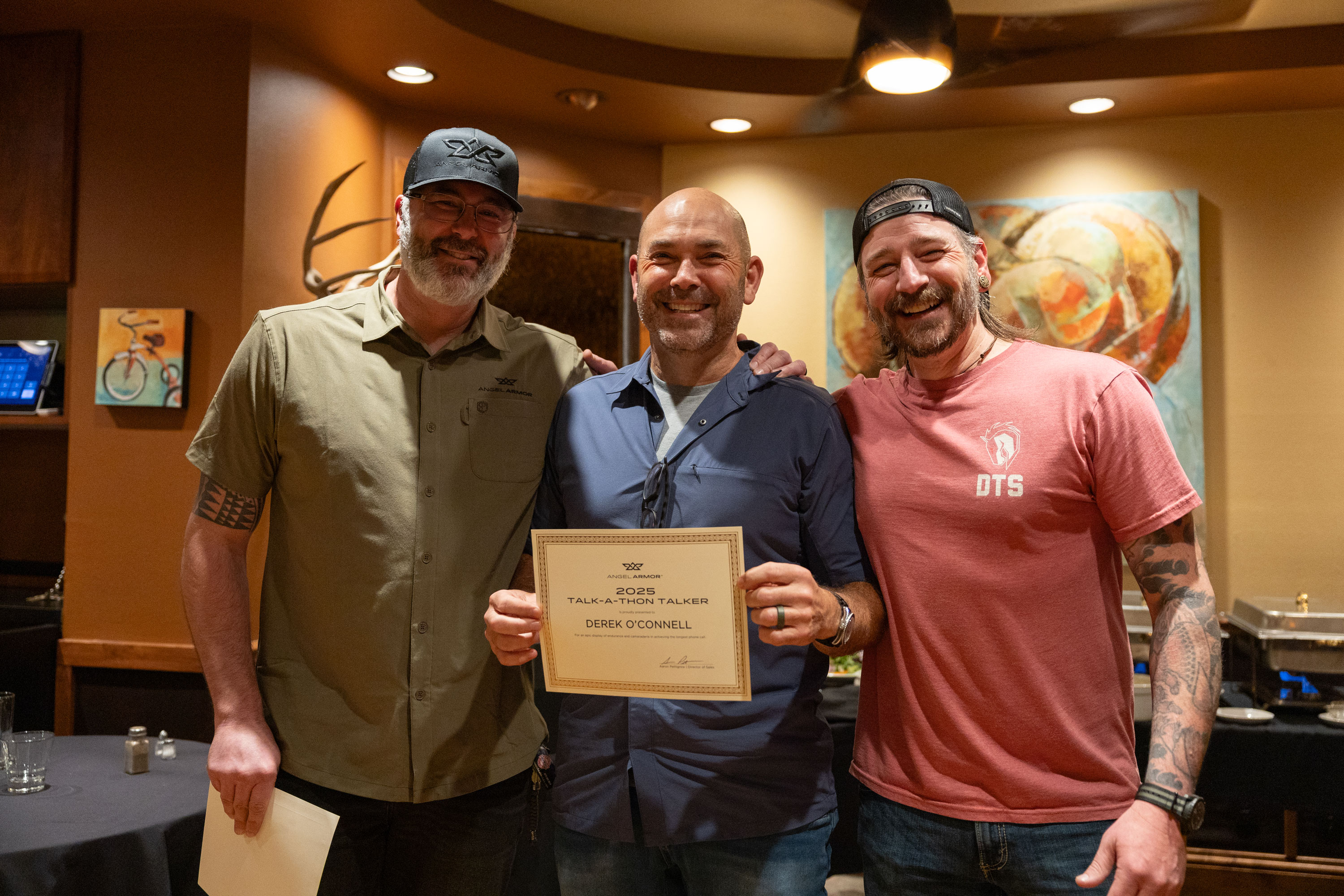Three men standing indoors smiling, one holding a certificate recognizing Derek O'Connell for Talk-A-Thon Talker 2025.