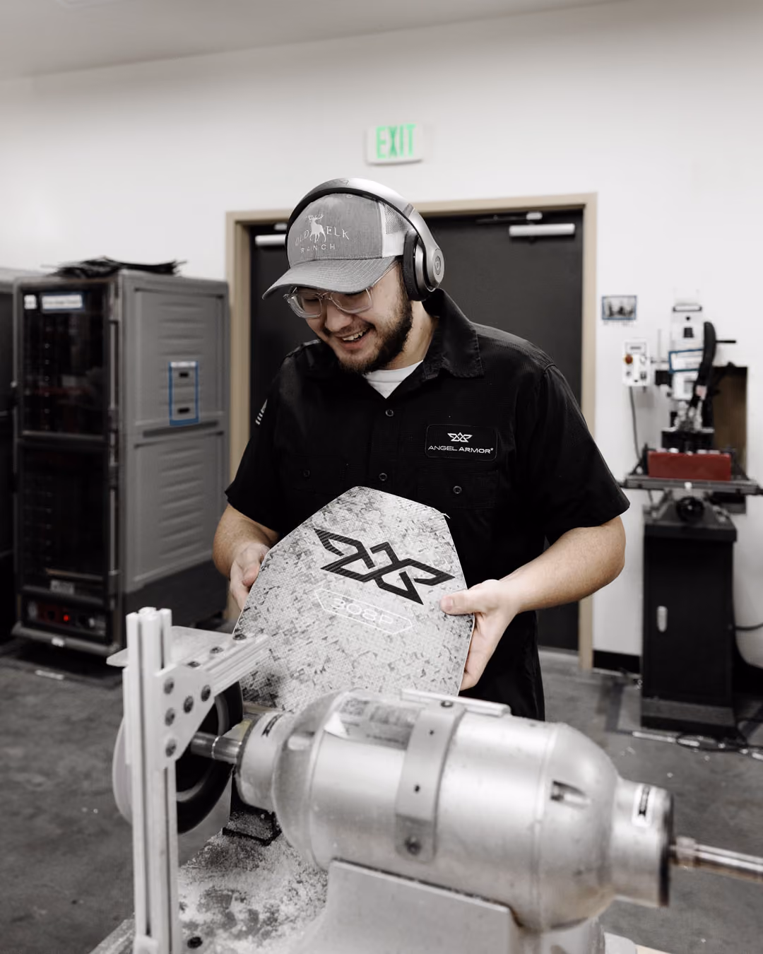 Man in a cap and headphones working on a piece of armor labeled 3082 in a workshop.