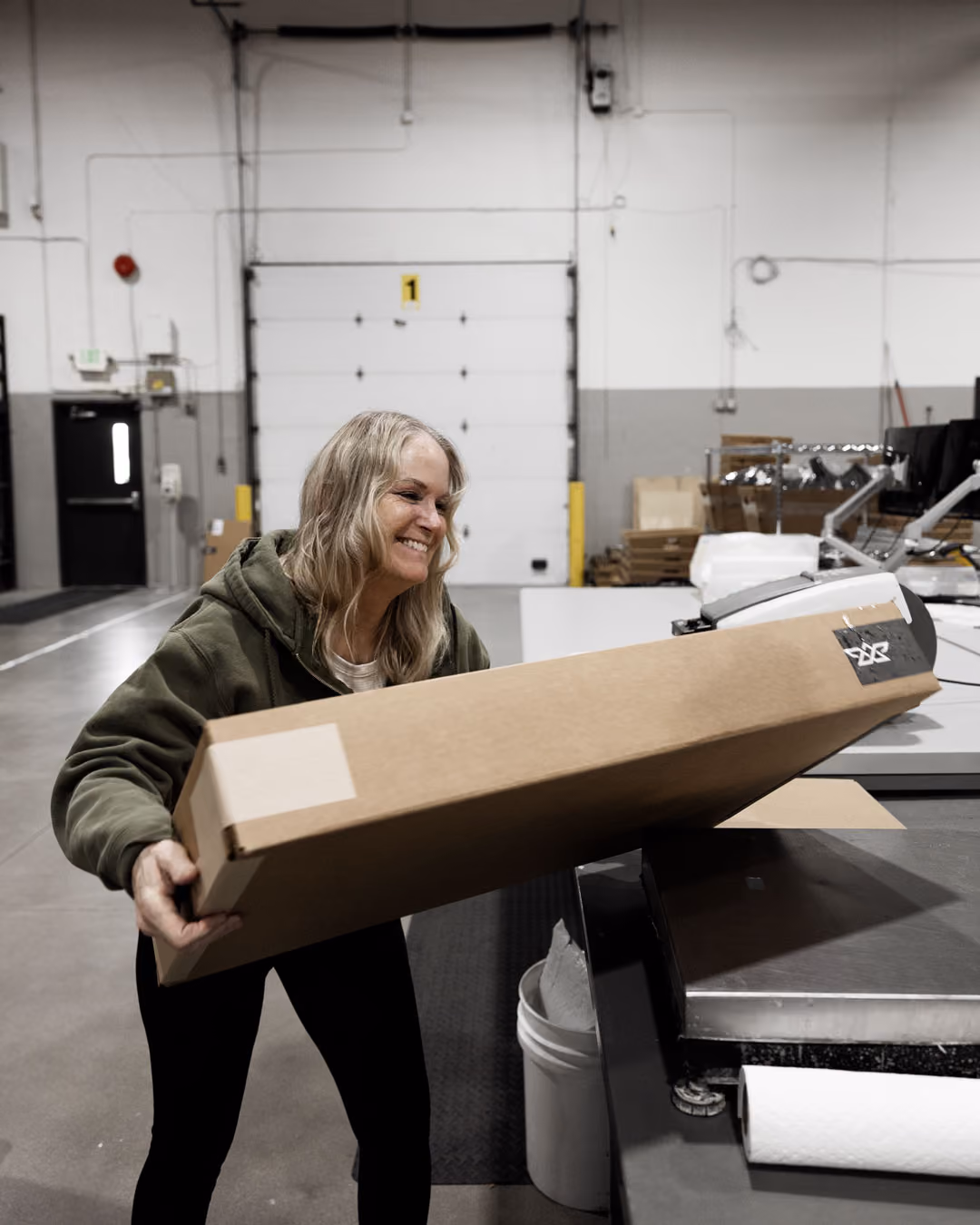 Smiling woman in green hoodie holding a large cardboard box in a warehouse setting.