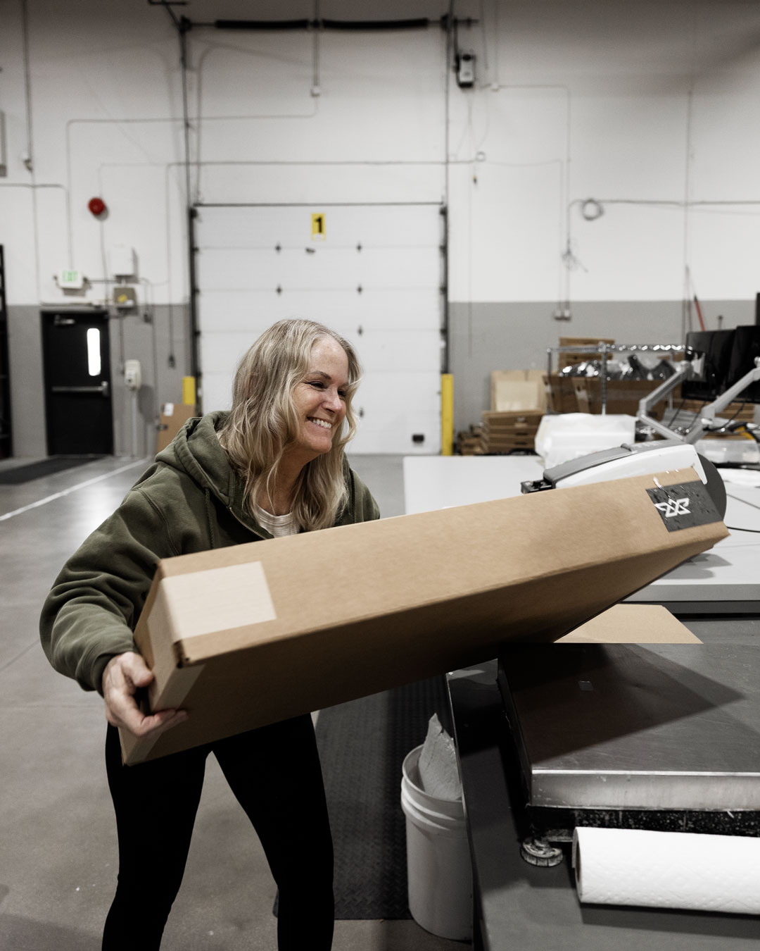 Smiling woman in green hoodie holding a large cardboard box in a warehouse setting.