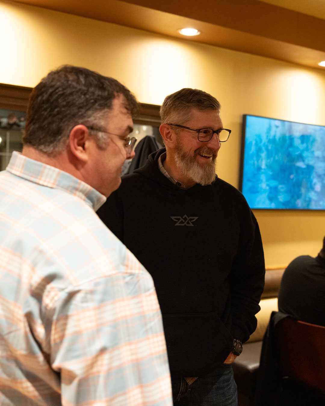 Two men in glasses chatting indoors, one in a plaid shirt and the other in a black hoodie.