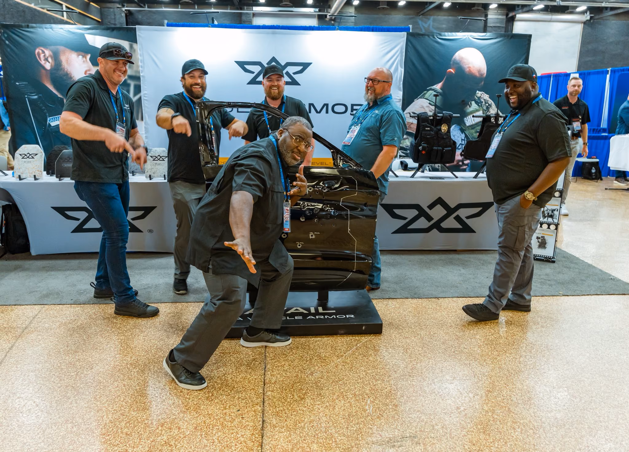 Group of men smiling and posing around a bulletproof car door at an exhibition booth.
