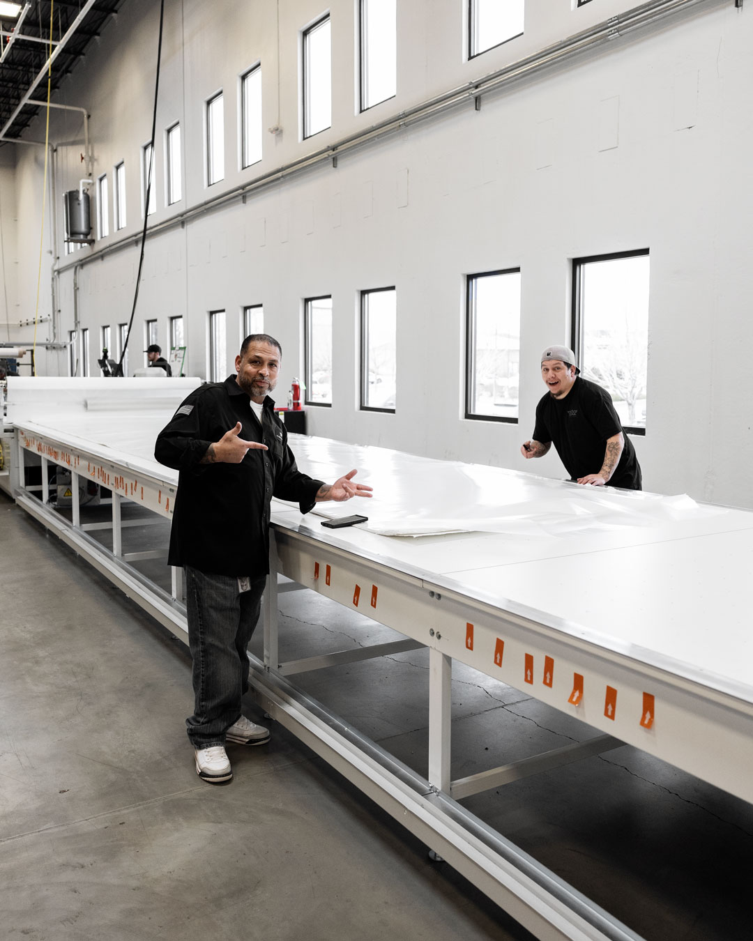 Two men working at a large flat table in a bright industrial workshop with rows of windows.