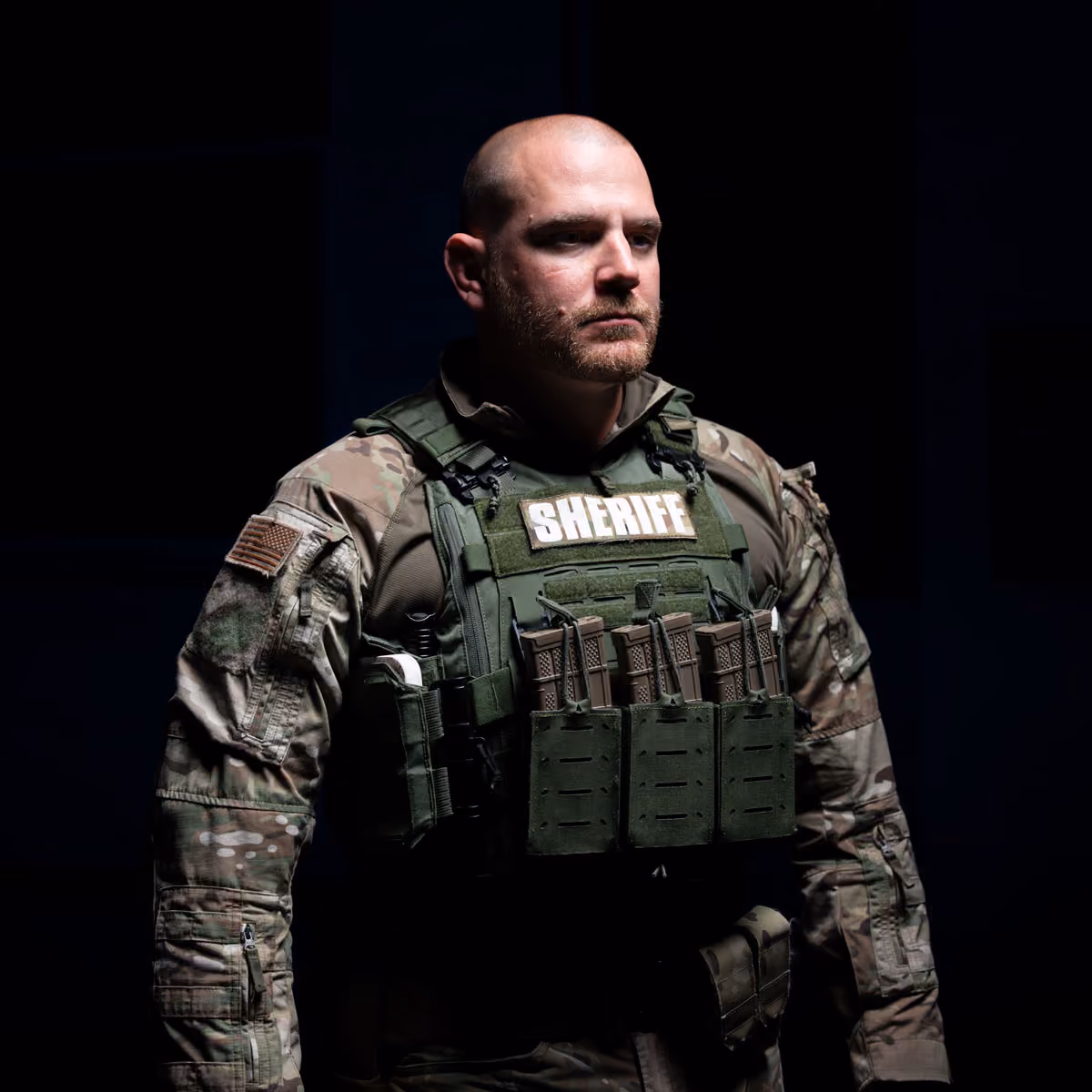 Serious male sheriff in tactical camo uniform and bulletproof vest with ammo magazines against black background.