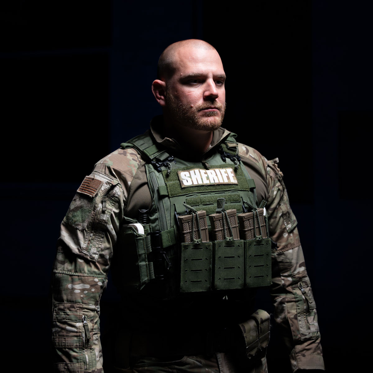 Serious male sheriff in tactical camo uniform and bulletproof vest with ammo magazines against black background.