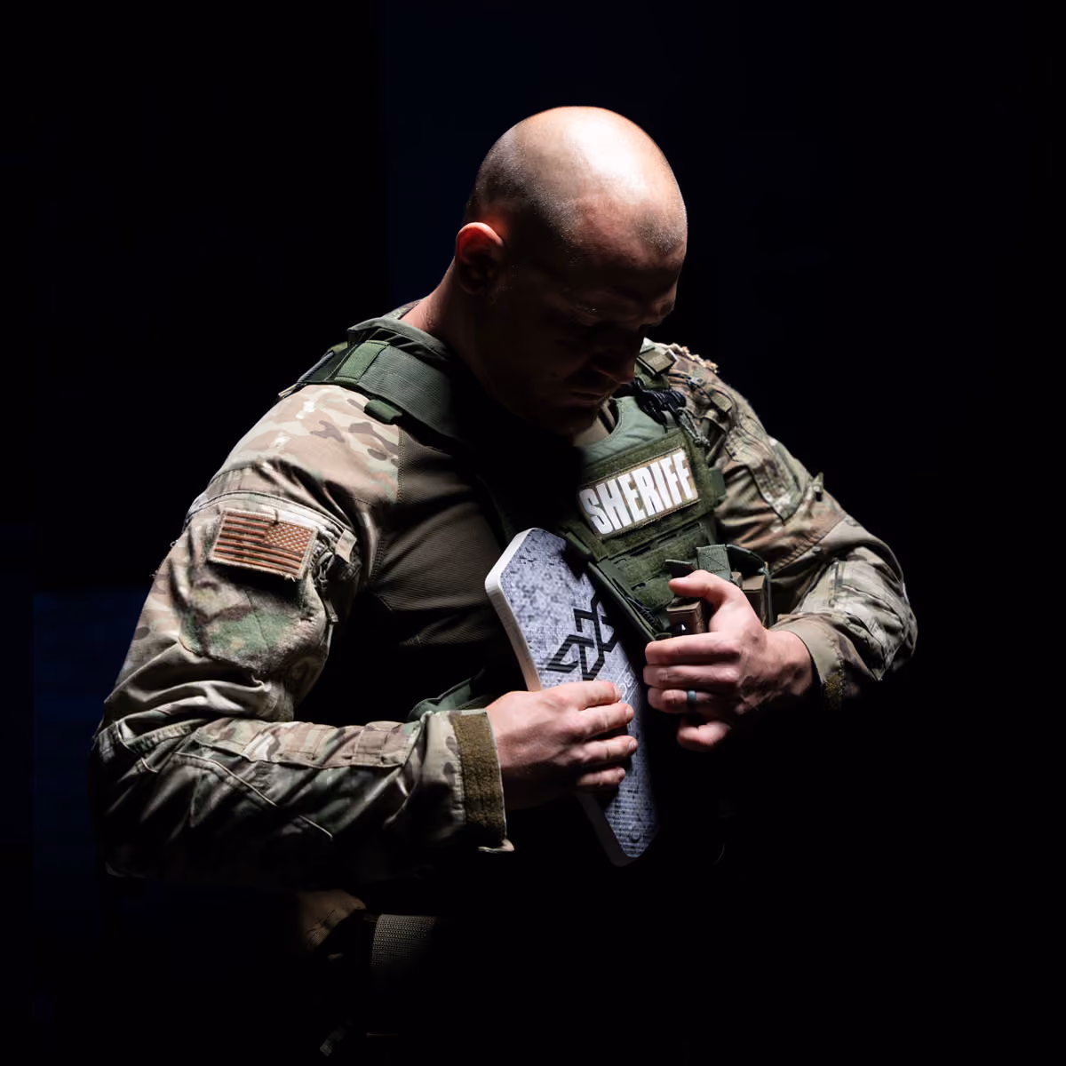 Bald sheriff in camouflage uniform holding a body armor plate against a dark background.