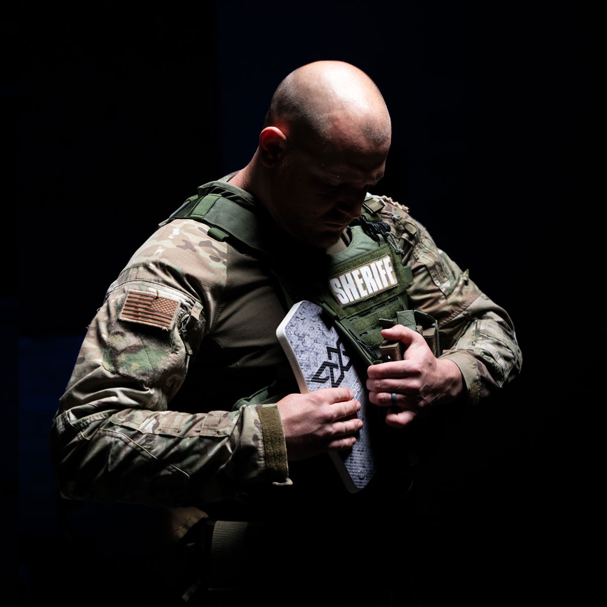 Bald sheriff in camouflage uniform holding a body armor plate against a dark background.