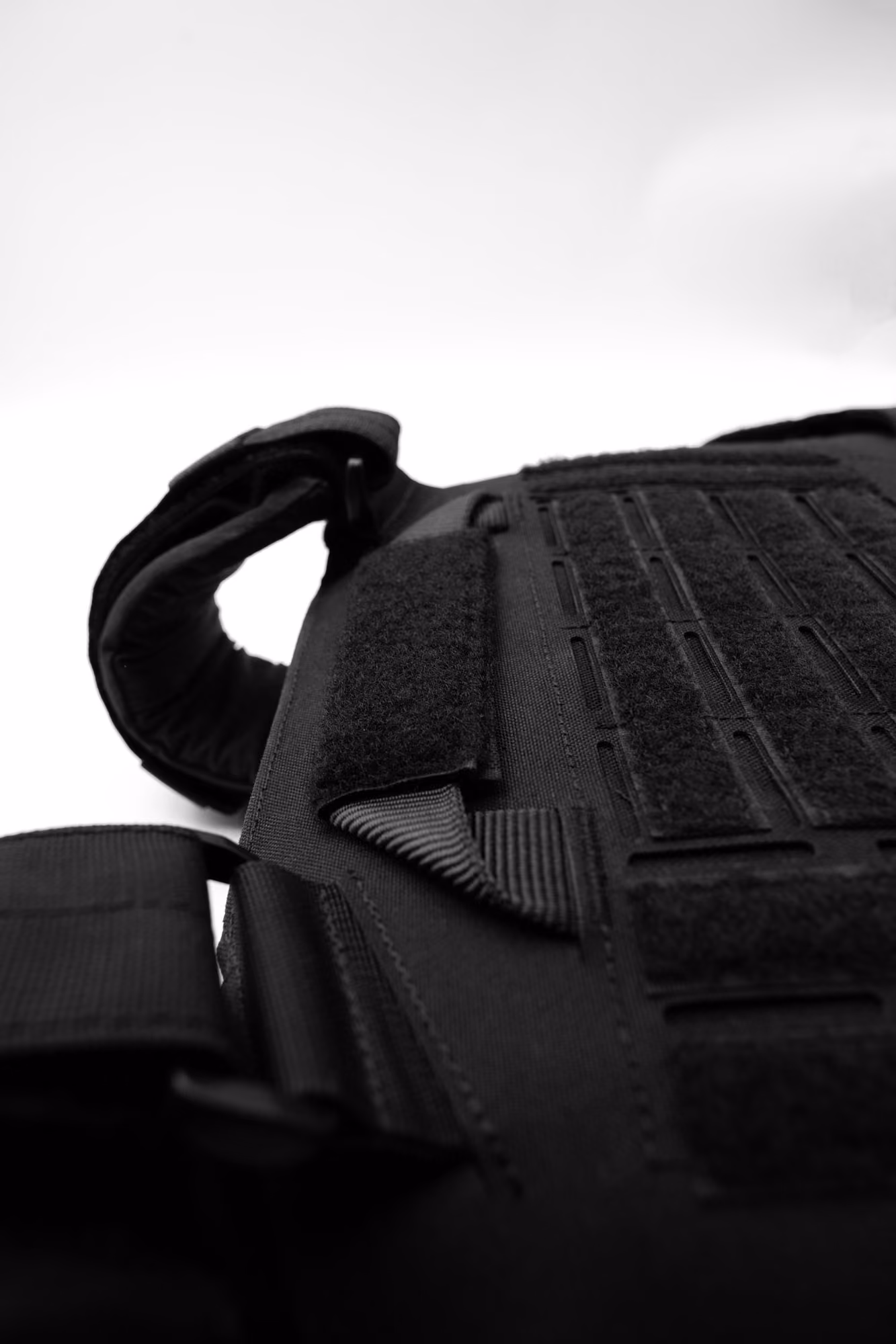 Close-up of a black tactical vest with velcro straps and padding on a white background.