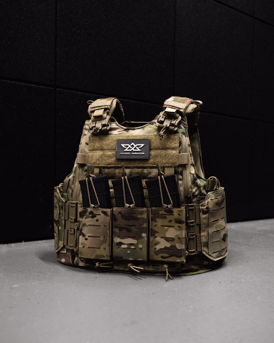 Camouflage tactical vest with four black rifle magazines on a gray floor against a black wall.