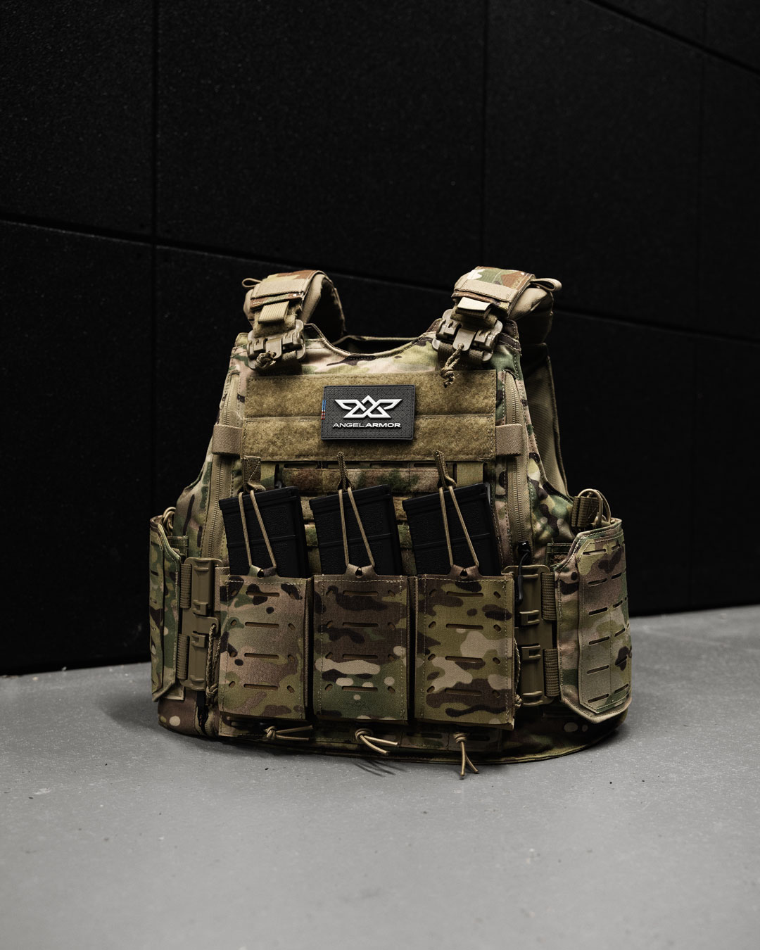 Camouflage tactical vest with four black rifle magazines on a gray floor against a black wall.