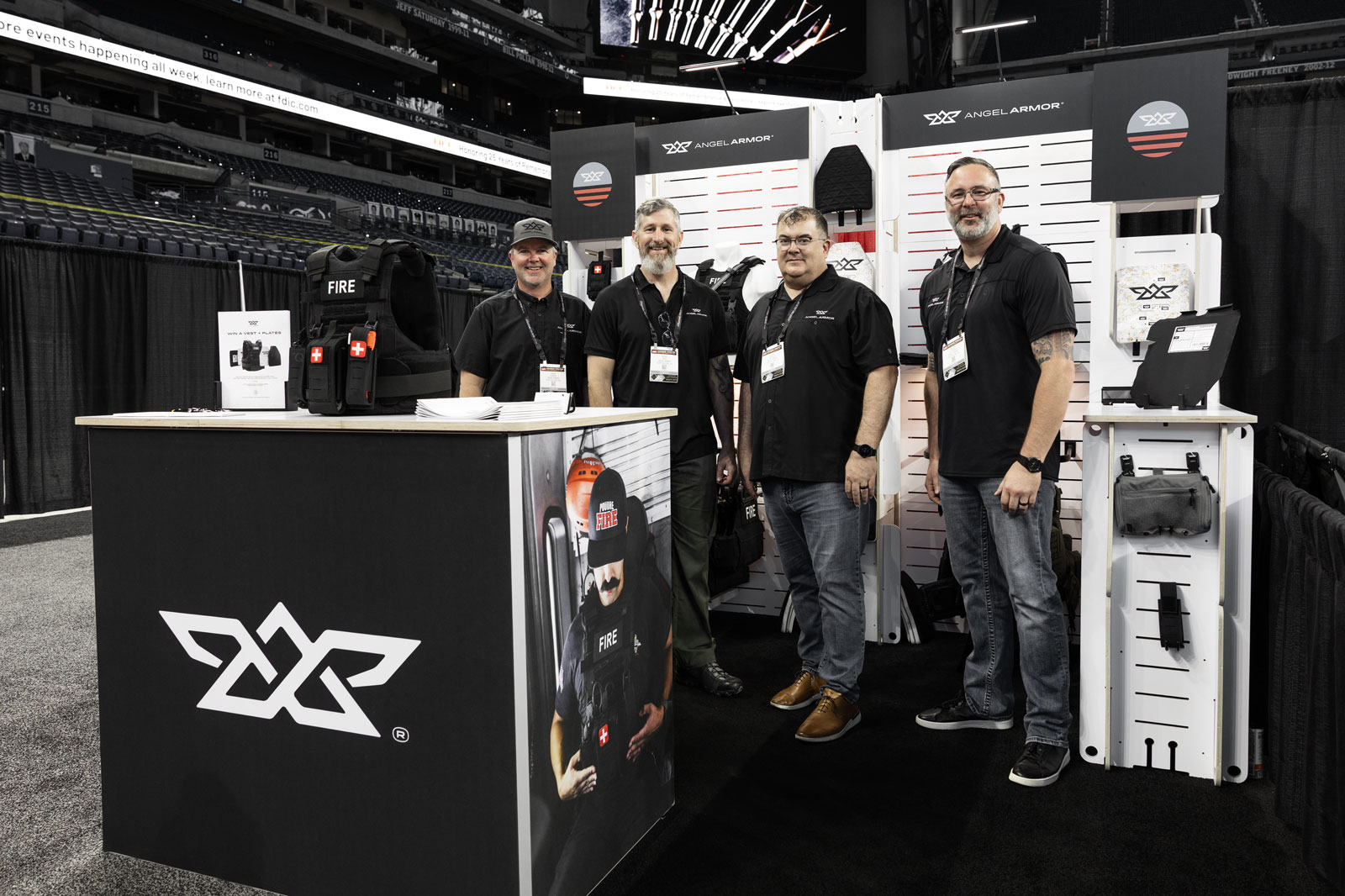 Four men in black shirts standing at an Angel Armor booth with tactical gear displayed.