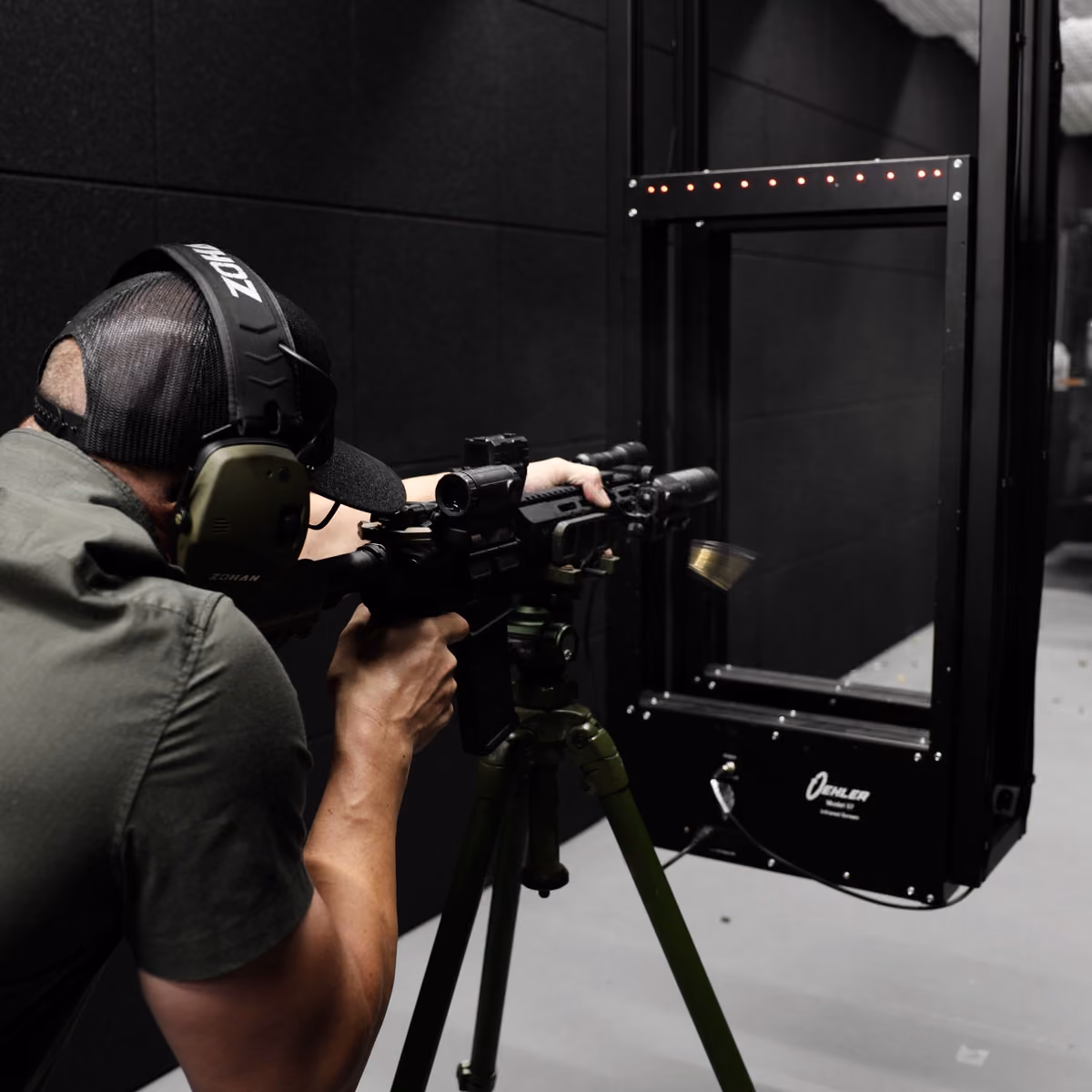 Man wearing hearing protection shooting a rifle mounted on a tripod at an indoor shooting range.