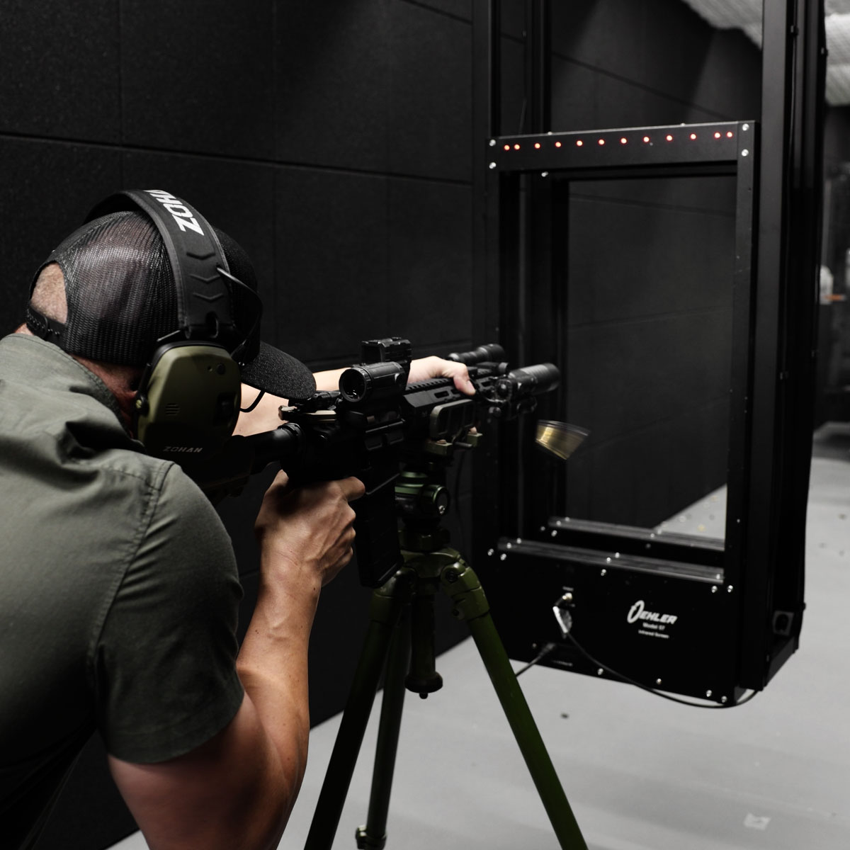 Man wearing hearing protection shooting a rifle mounted on a tripod at an indoor shooting range.