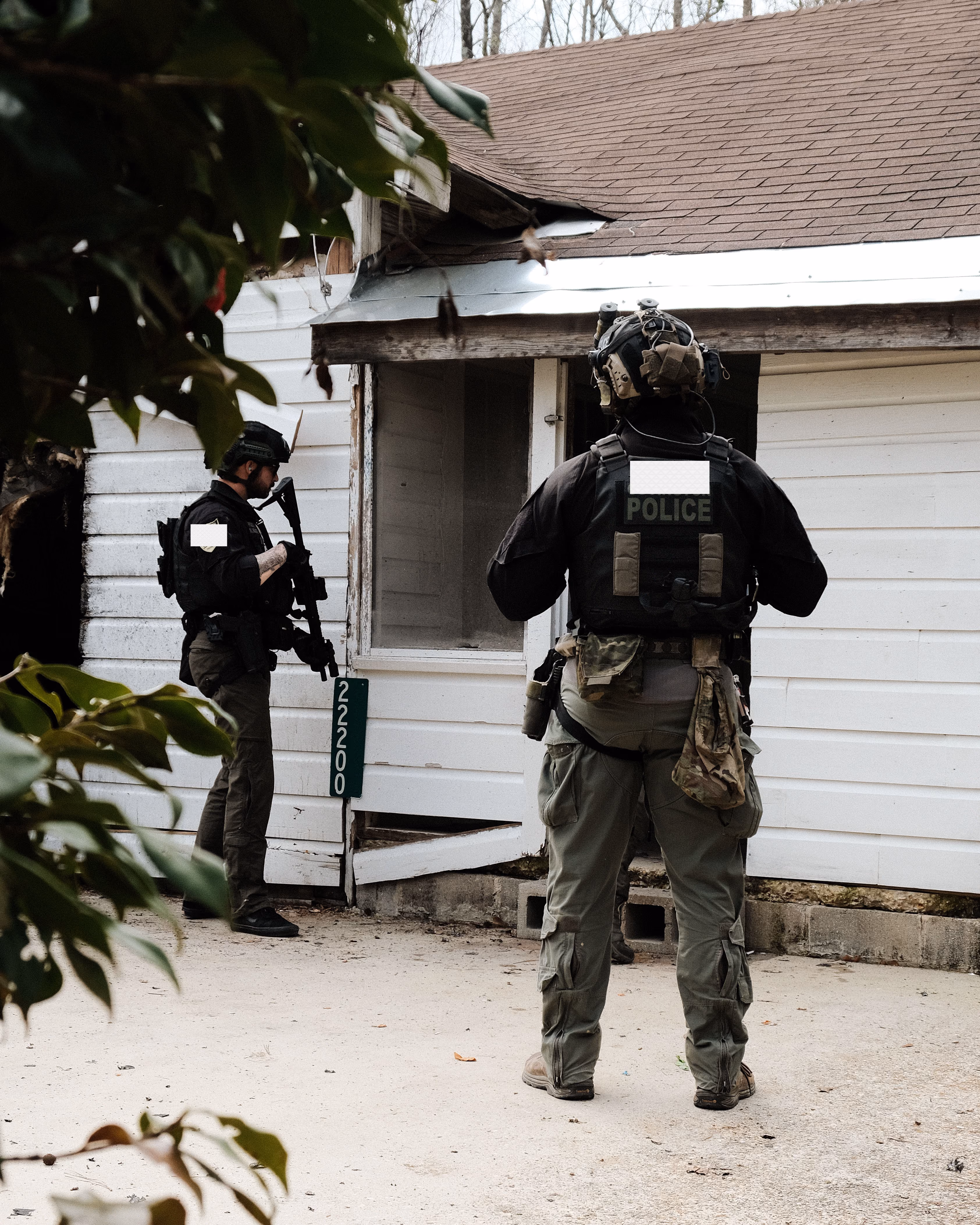 Two armed police officers in tactical gear stand outside a white house with damaged siding near address 22200.