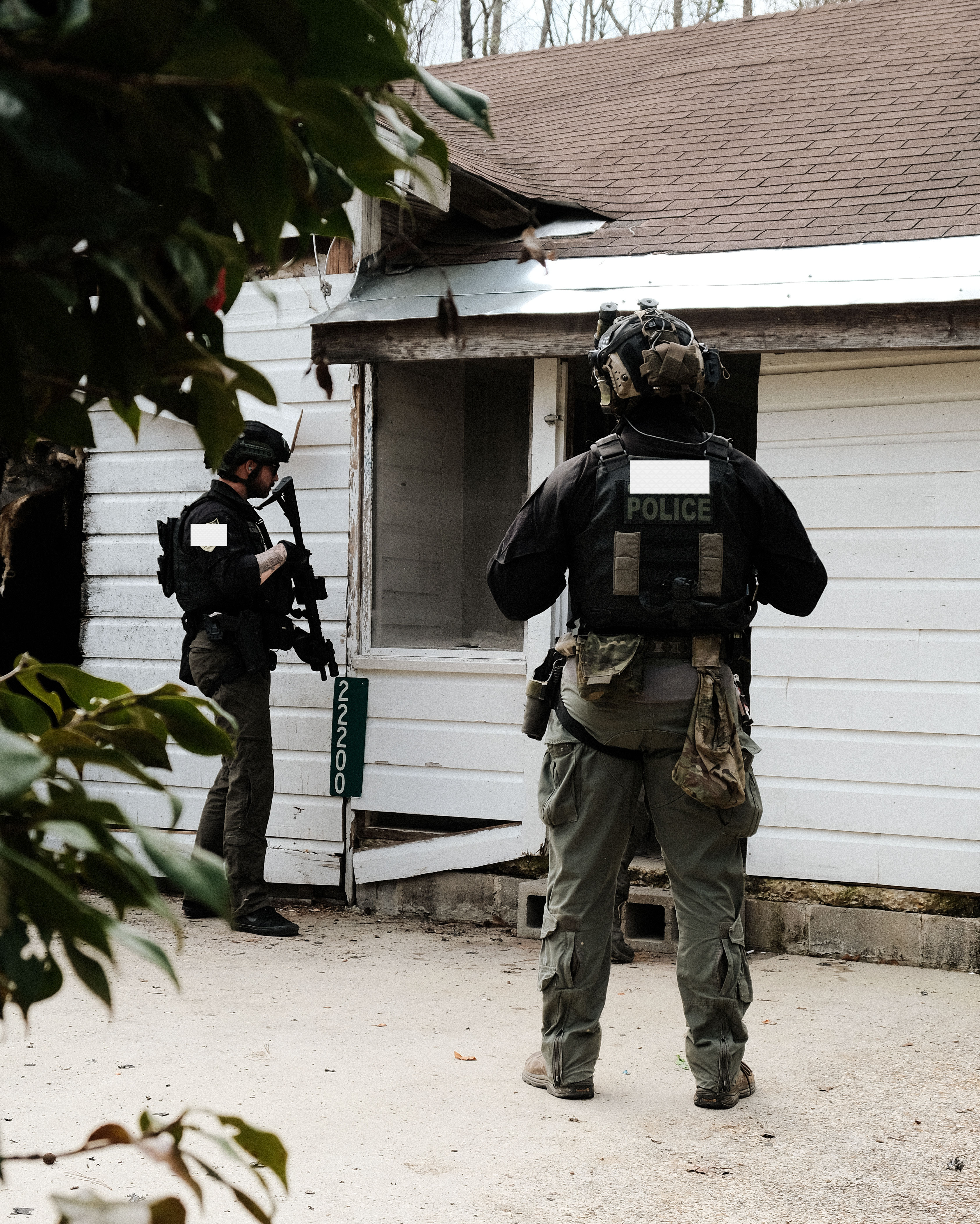 Two armed police officers in tactical gear stand outside a white house with damaged siding near address 22200.