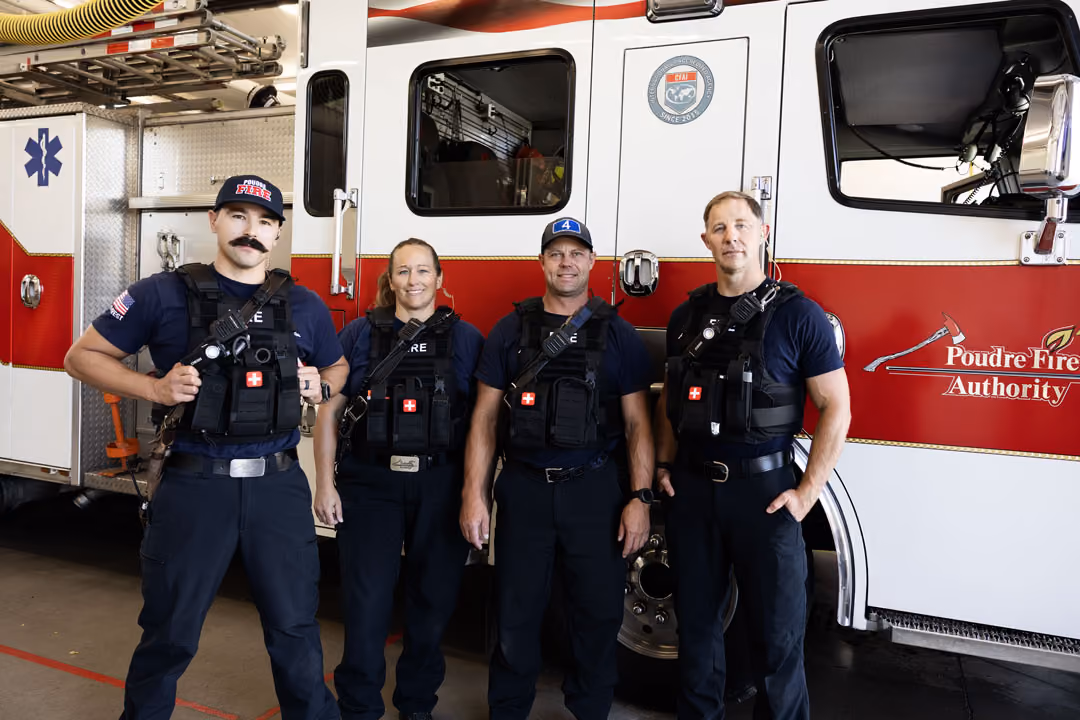 Four firefighters in navy uniforms and gear standing in front of a red and white fire truck.