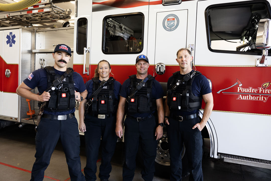 Four firefighters in navy uniforms and gear standing in front of a red and white fire truck.