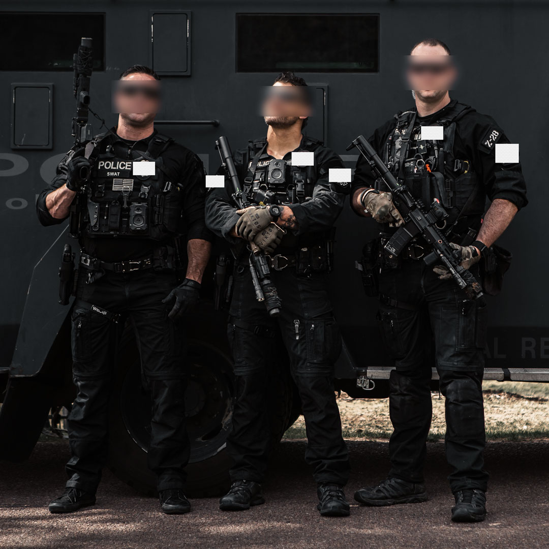 Three armed SWAT officers in black tactical gear standing in front of an armored vehicle.