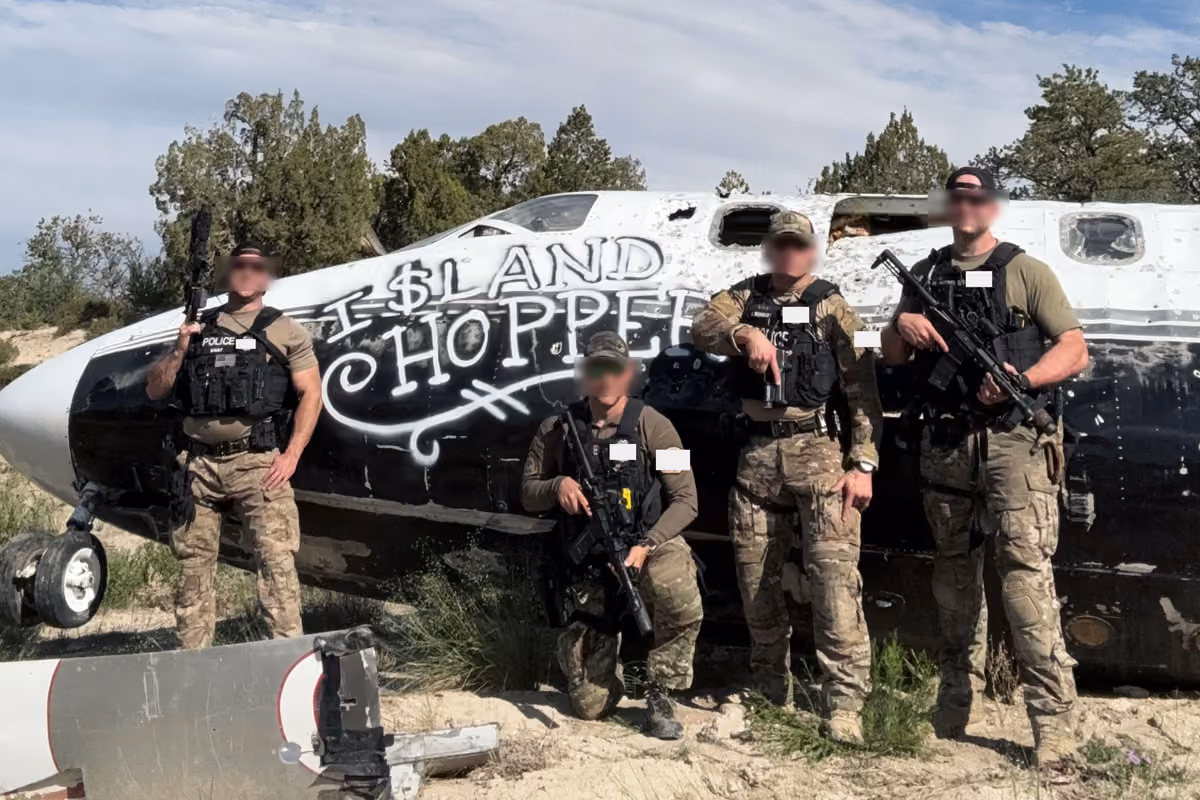 Four armed police officers in tactical gear posing in front of a damaged black-and-white airplane.