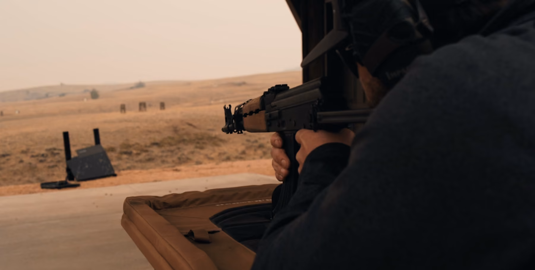 Person aiming a rifle at a shooting range with targets in a barren landscape.