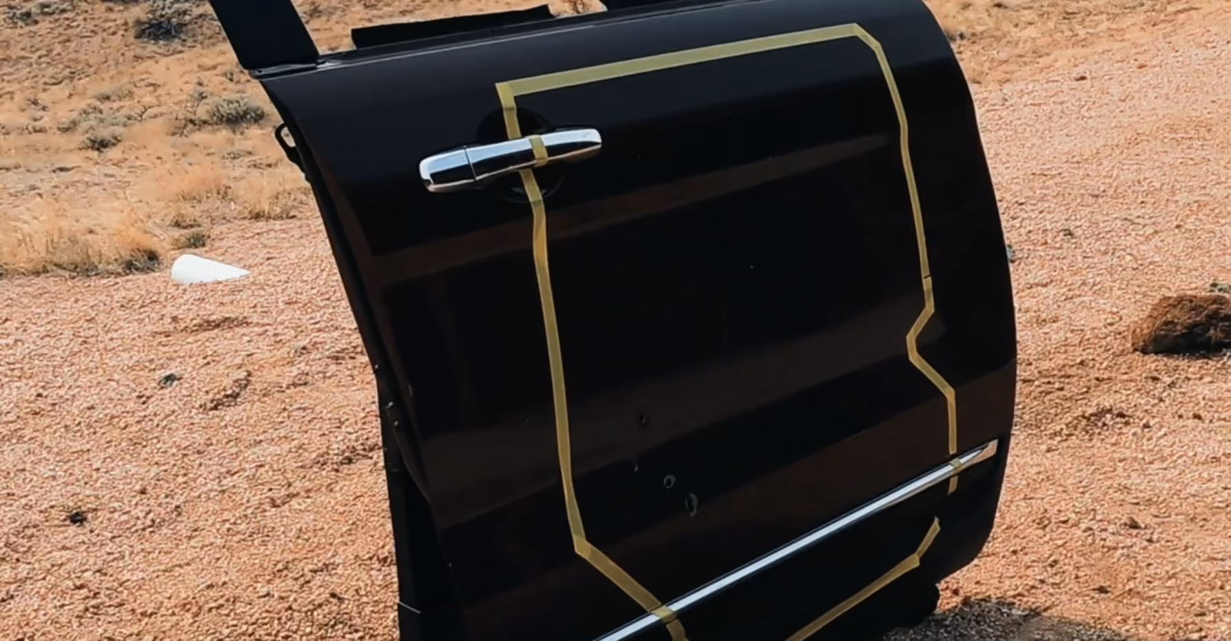 Black car door with yellow tape outlining dents and damage on a gravel surface outdoors.