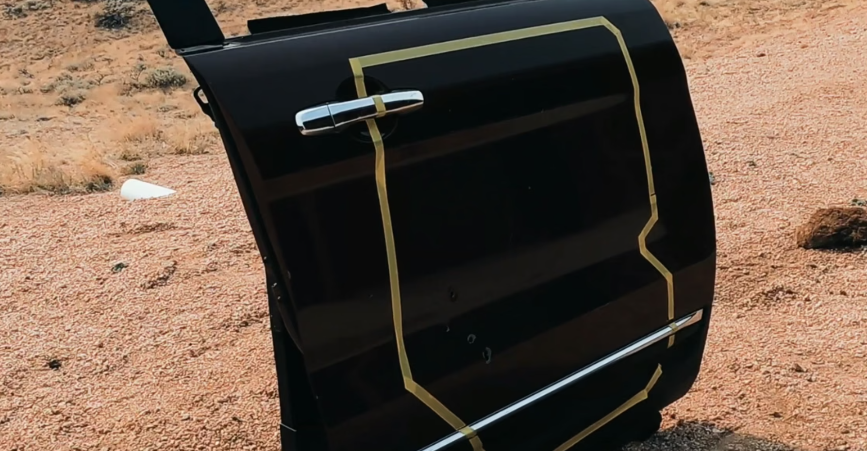 Black car door with yellow tape outlining dents and damage on a gravel surface outdoors.