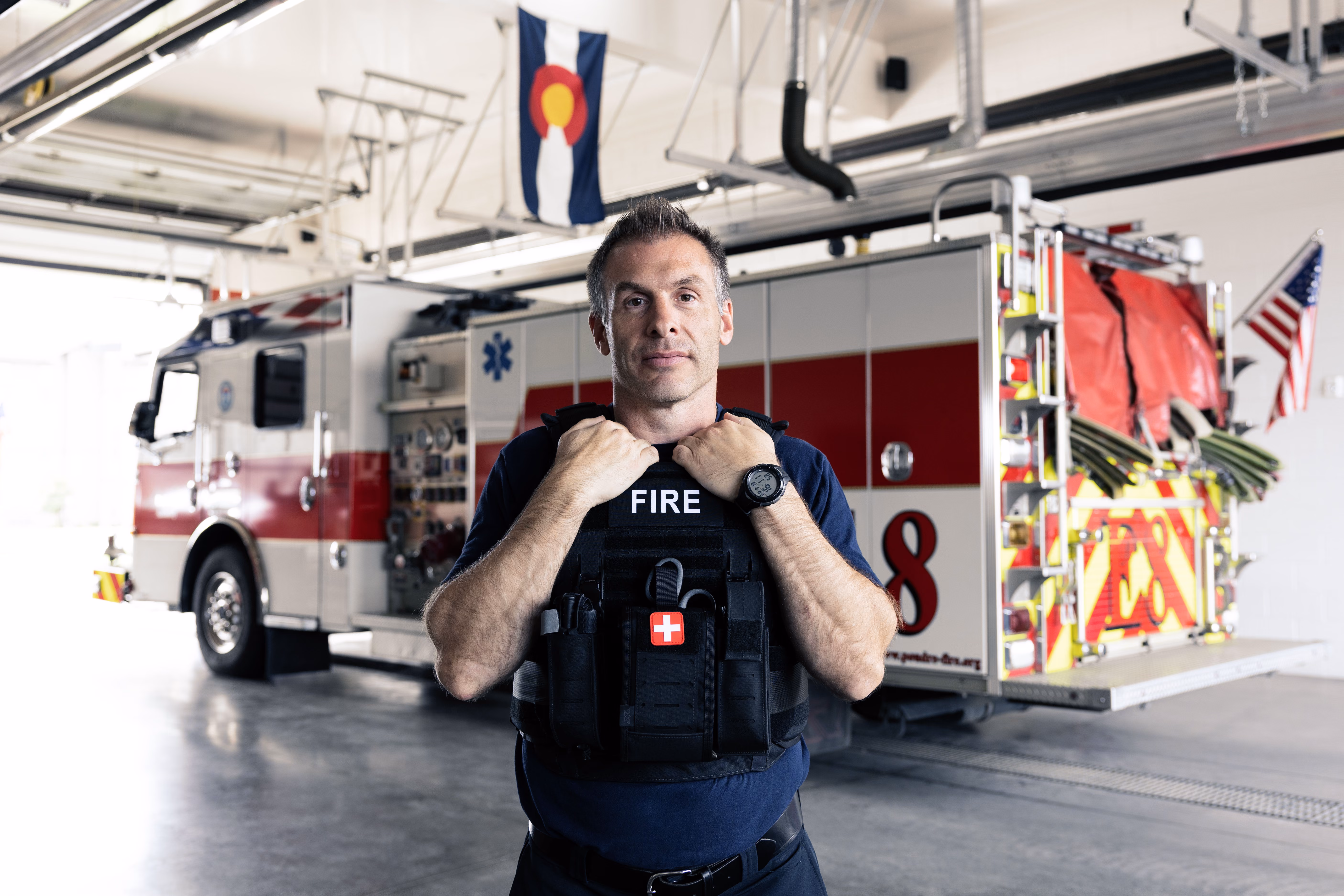 Firefighter wearing a black vest standing in a fire station with a fire truck and flags behind him.