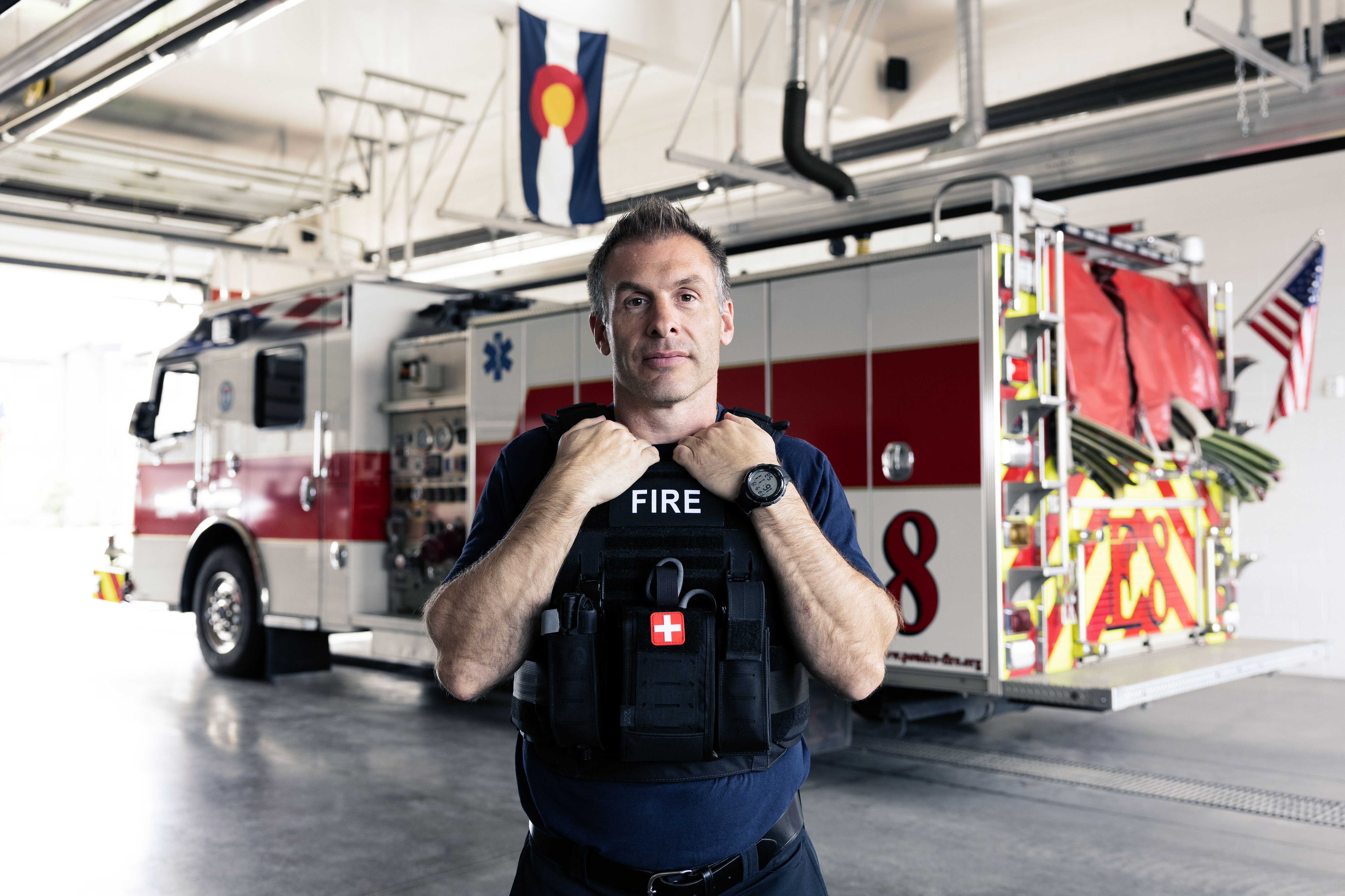 Firefighter wearing a black vest standing in a fire station with a fire truck and flags behind him.