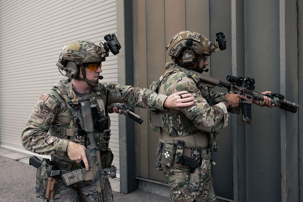 A soldier in camouflage uniform holding a tactical helmet and green body armor in low light.