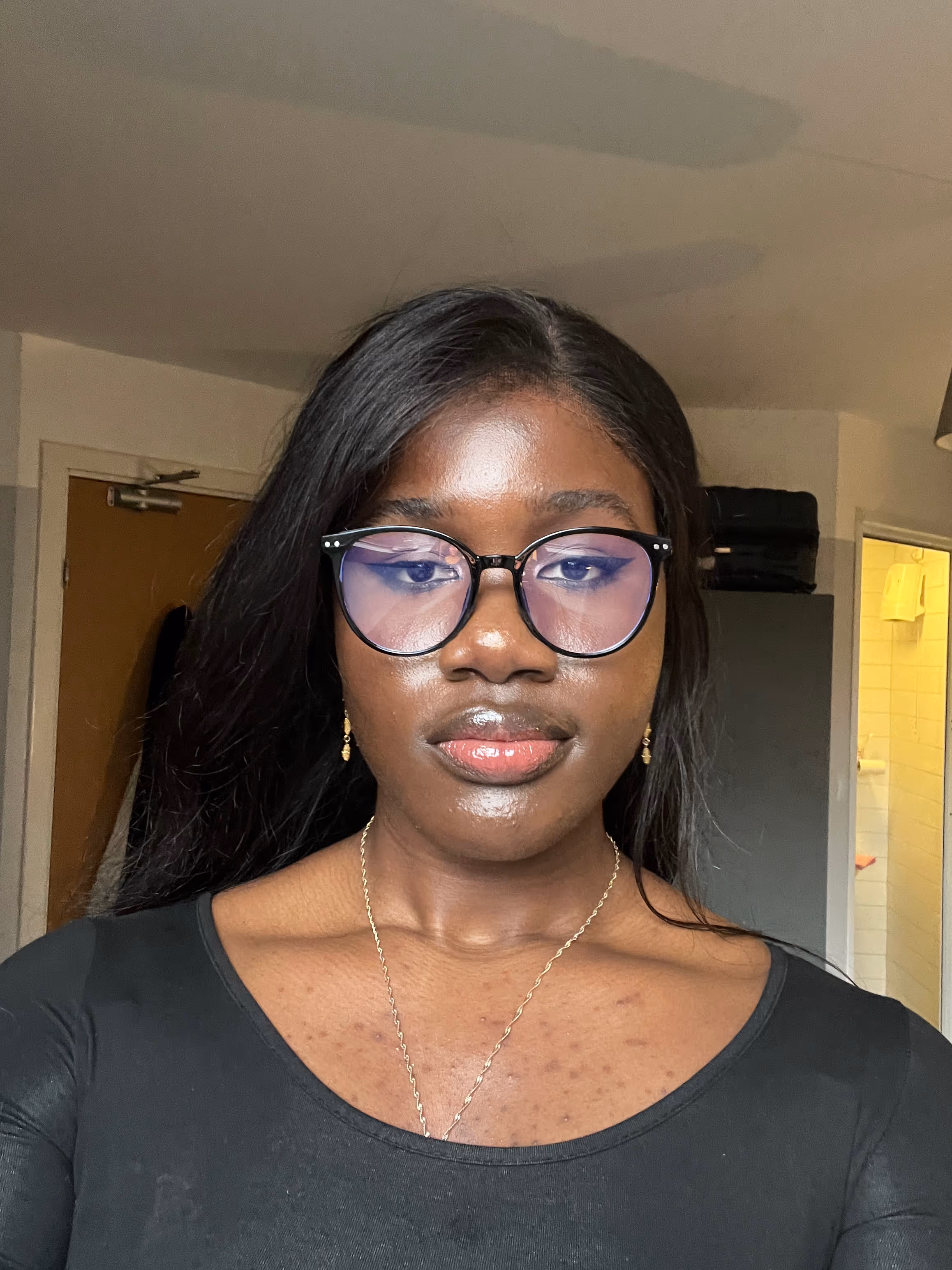 Woman with long black hair, wearing round glasses, gold earrings, a gold necklace, and a black top, standing indoors.