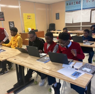 A classroom of students using laptops at long wooden desks