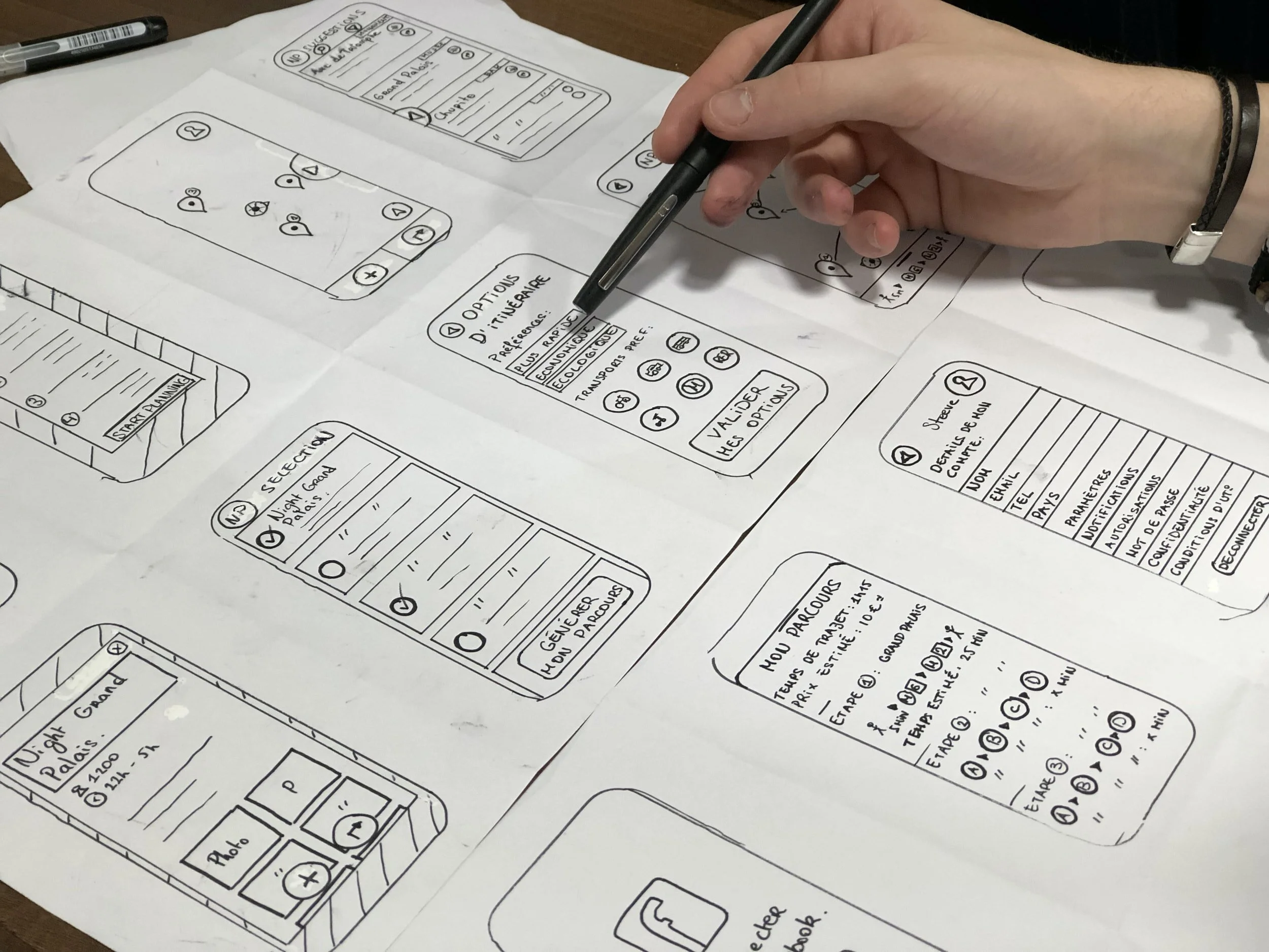 A hand and pen hovering over a large white paper sheet of mobile screen wireframe designs