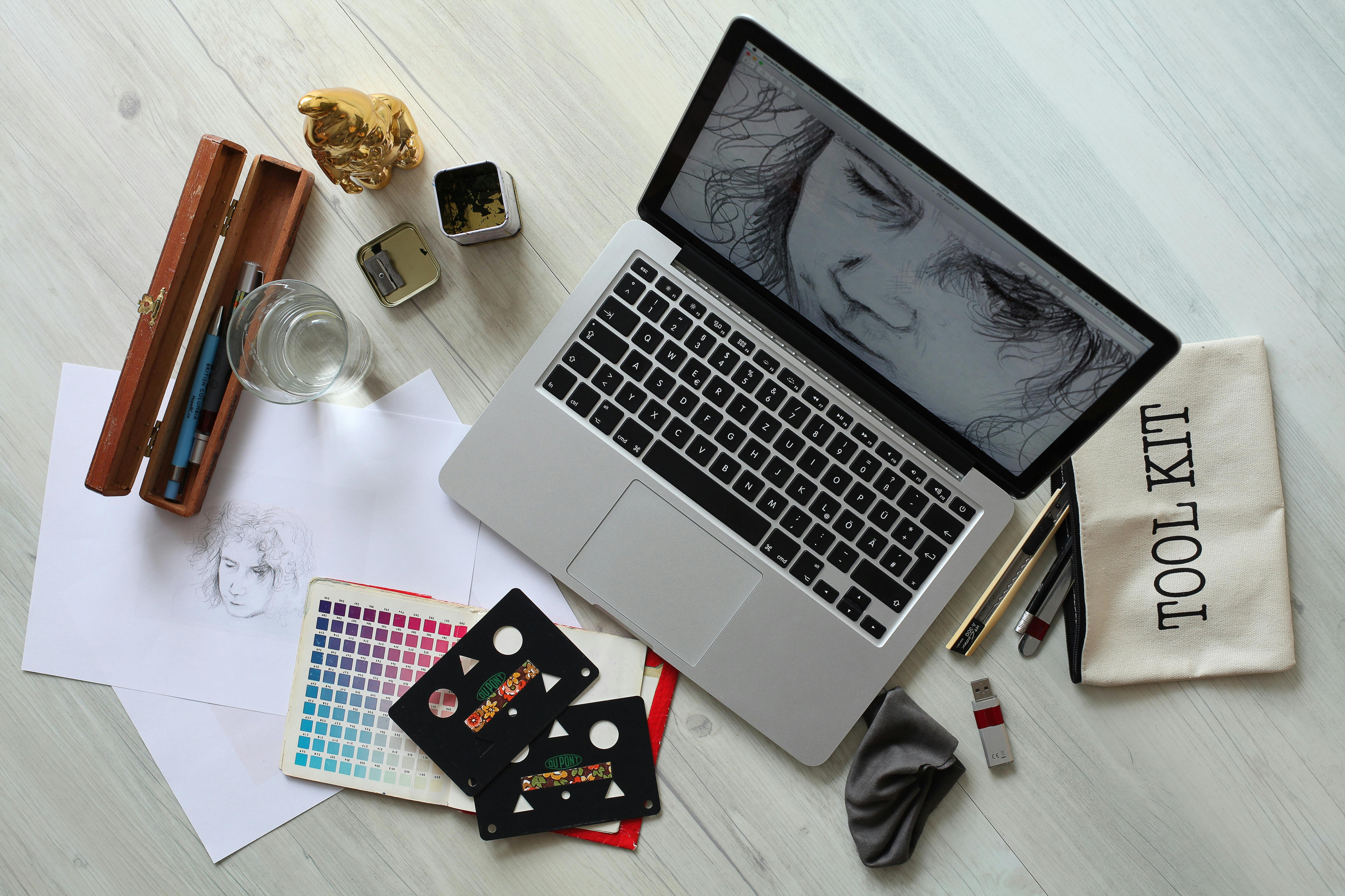 A top down view of a laptop screen with a pencil drawing of a face. Art supplies surround the computer and a pouch reads "toolkit."