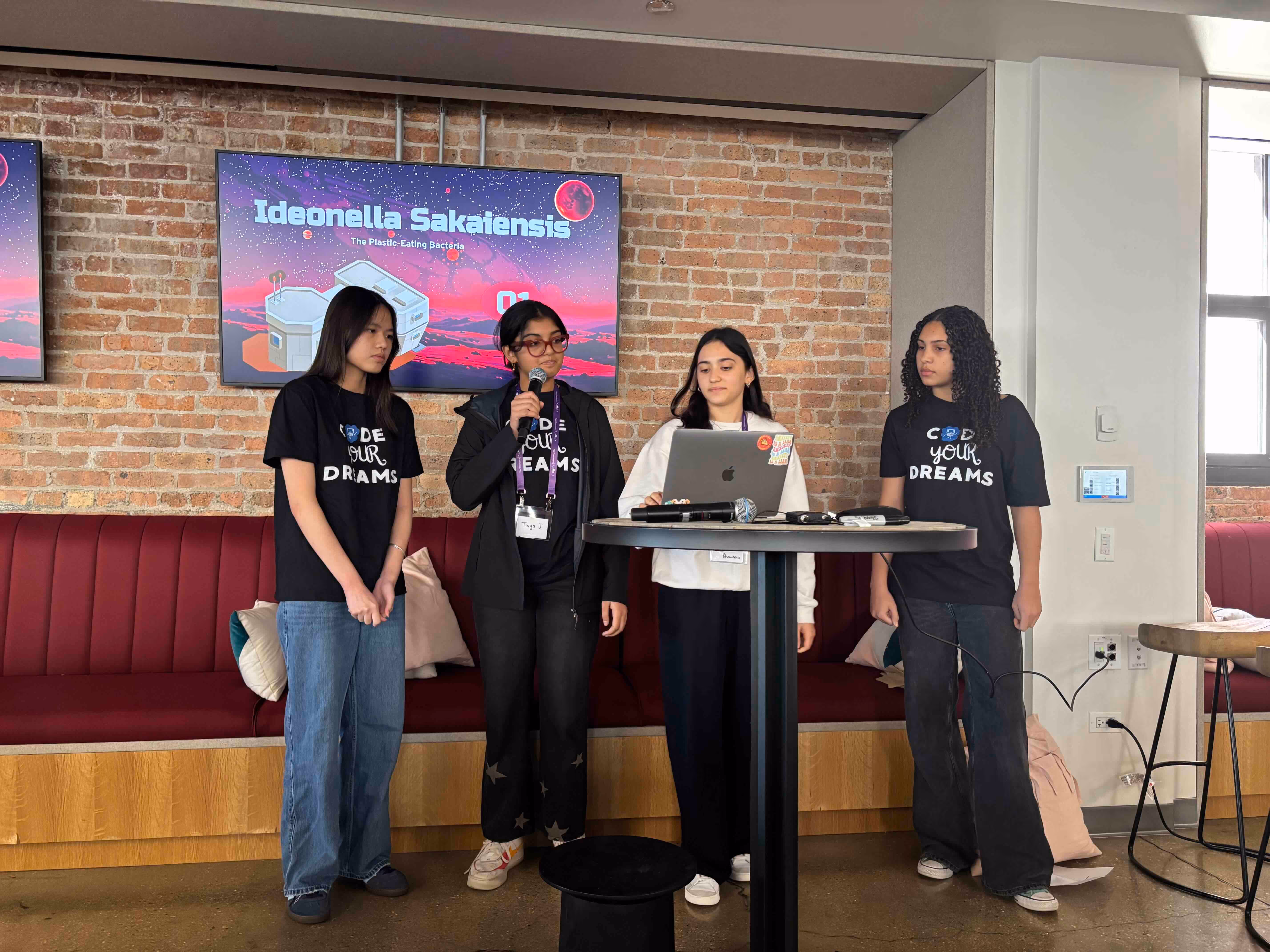 Four students stand at a tall table with a laptop on it. A sign behind them reads "Ideonella Sakaiensis, the plastic eating bacteria"