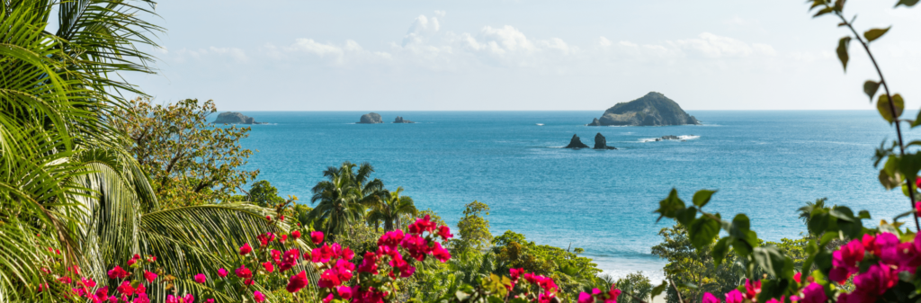 A view of the Costa Rican coast, framed by bright pink flowers and tropical foliage. The clear blue ocean stretches out to the horizon, dotted with several small, tree-covered islands.